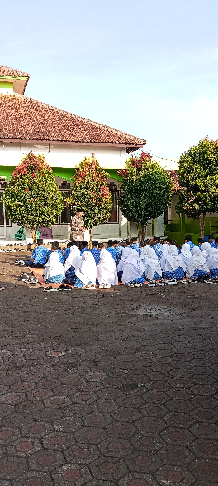 A group of people are sitting on the ground in front of a building in Cangkuang, Indonesia. The people are wearing white headscarves and blue shirts. They are sitting in rows, facing a man standing in front of them. The man is wearing a white headscarf and a blue shirt. There are trees in the background. The ground is paved with hexagonal tiles.