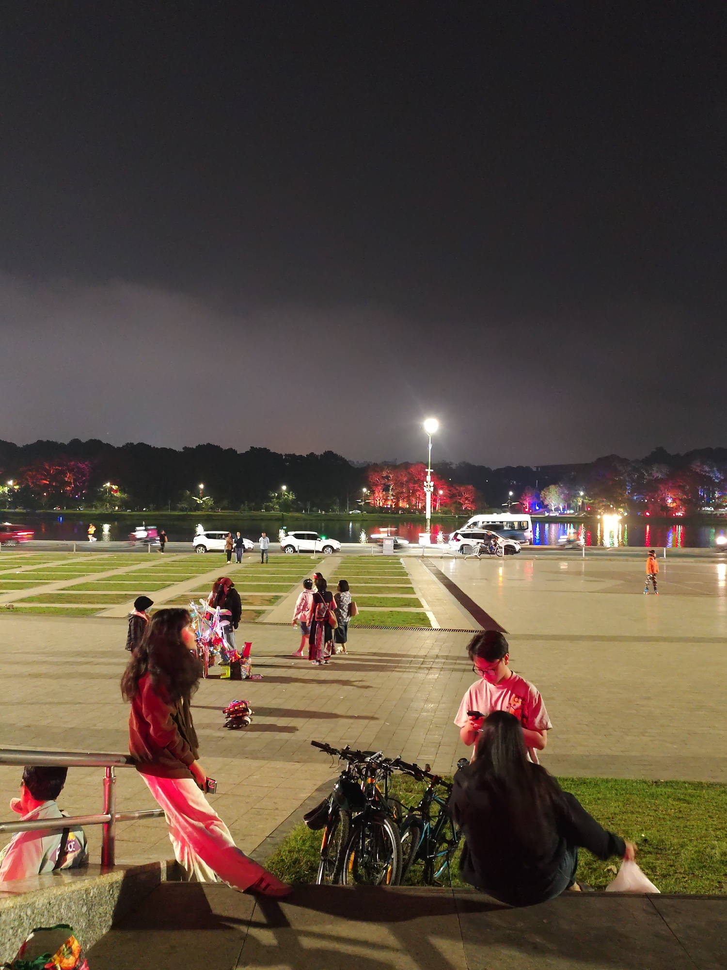 At night in Da Lat, Vietnam, a well-lit plaza fronts a body of water. The water reflects the city lights on the far side, where a line of trees defines the horizon. In the plaza, several people stand or walk, including a group around a street vendor with colorful wares. In the foreground, a girl leans on a metal railing, while another person sits on a step next to her. Two bicycles are parked nearby. To the right, a boy stands using a phone, facing a seated person with long hair. The sky is dark and cloudy.
