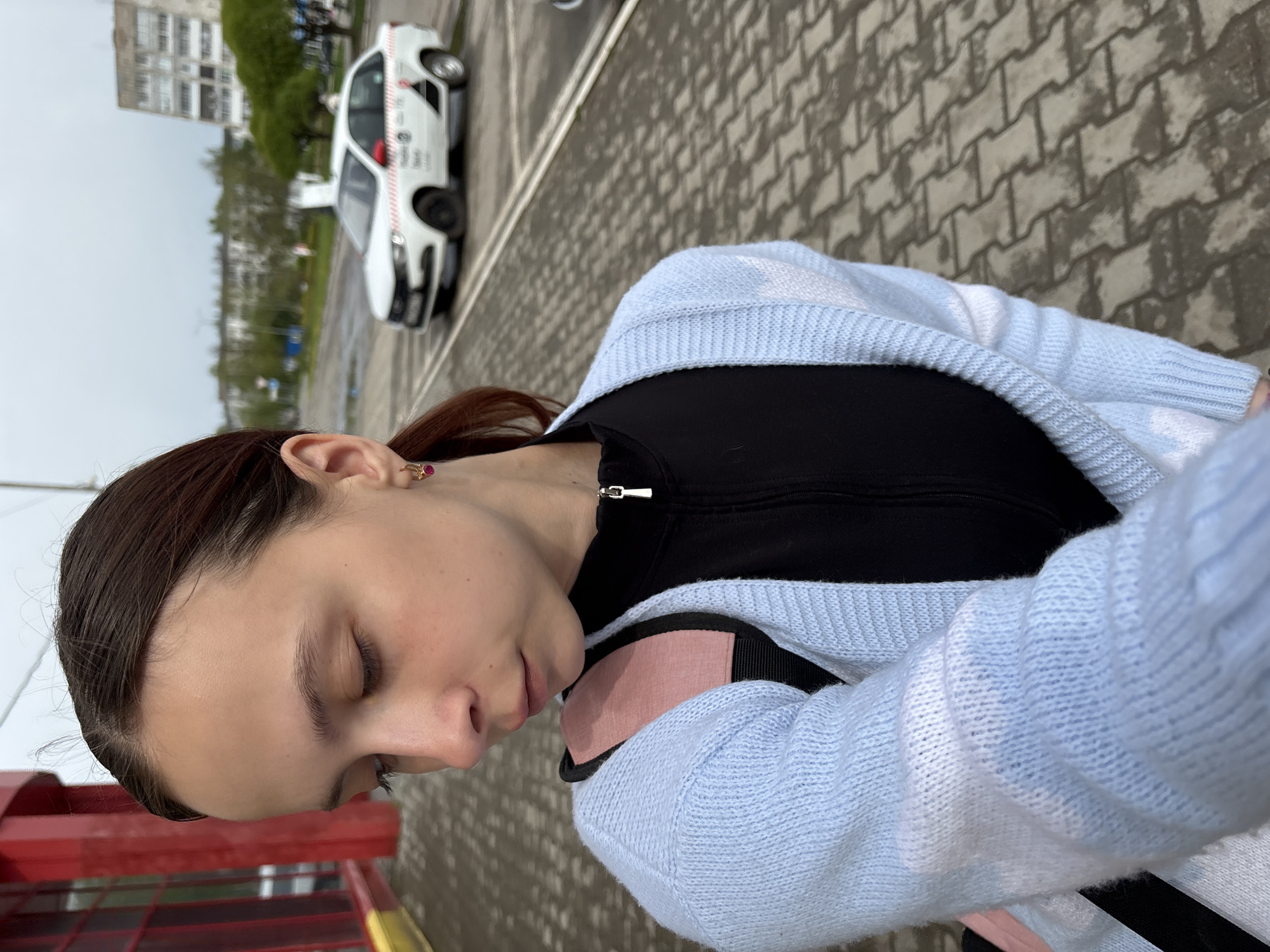 A young woman with brown hair is looking down in a close-up selfie. She has a black zippered top underneath a light blue cardigan with white stars. She's wearing an earring in her left ear.
Behind her, out of focus, there's a parking area. A white vehicle with red and black markings is visible, and a multi-story building is in the background. The image appears to be taken in Chaikovsky, Russia.