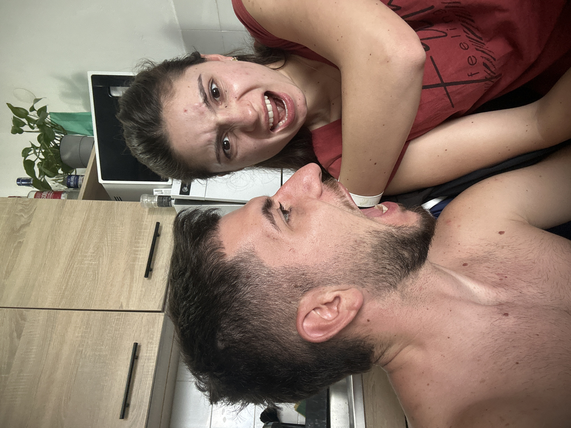 The image shows a man and a woman posing for a photograph. The woman is wearing a red t-shirt with a black design. The man is shirtless and has a beard. Both have their mouths open as if screaming. The setting appears to be a domestic bathroom. Behind the woman, there is a small cabinet with a green plant and some bottles of liquid. Behind the man, there are wooden cabinets. There is no information to suggest the location is Sofia, Bulgaria.