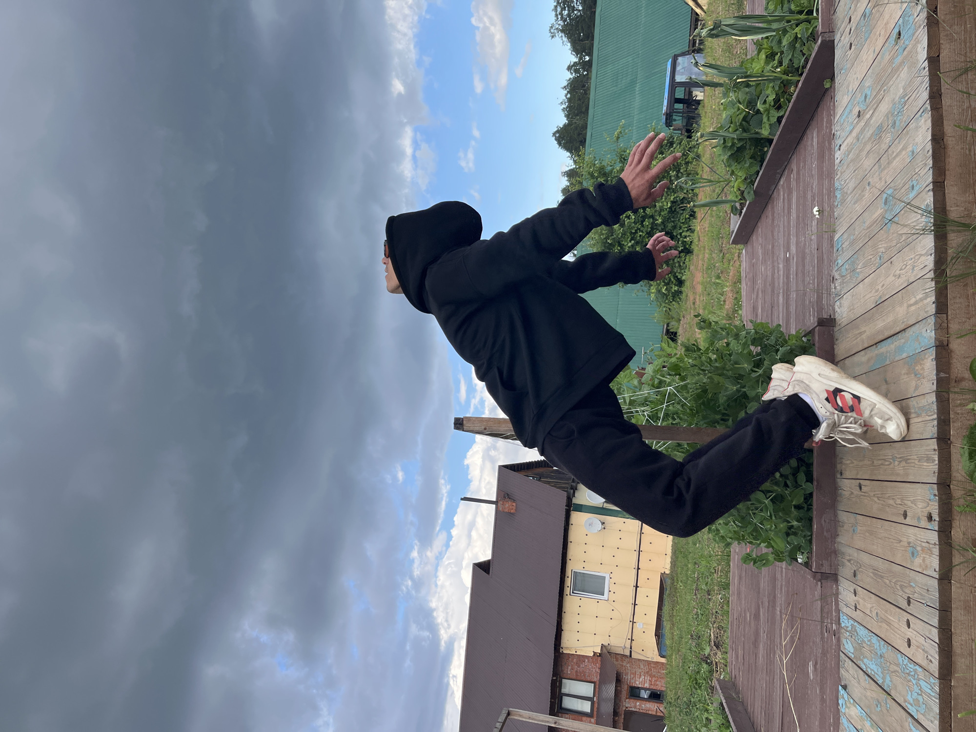 The image captures a person in mid-air, suspended horizontally with their head tilted back, wearing a black hoodie and black pants with white sneakers. They are positioned over a wooden structure that appears to be a raised garden bed with plants growing in it. In the background, there is a house with a brown roof and yellow siding, and another building with a green roof. The sky is cloudy with patches of blue. The scene appears to be in a rural or suburban setting, likely a backyard or garden area, possibly in Chaikovsky, Russia, given the architectural style of the buildings and the presence of what looks like a dacha (summer house). The overall impression is one of movement and dynamic posing against an outdoor backdrop.