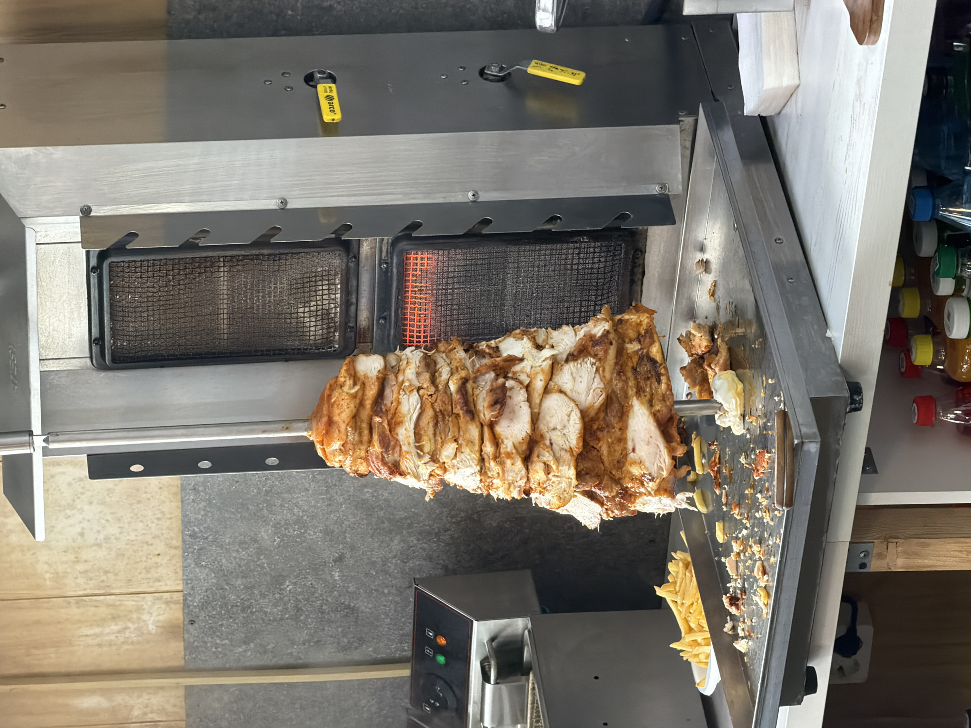 The image shows a vertical rotisserie kebab machine with a partially carved cone of meat, likely chicken or a mix of meats, positioned in front of a gas-powered infrared broiler. The broiler has a safety shut-off valve with a yellow handle. A collection of shaved meat and French fries are visible on a work surface adjacent to the rotisserie, suggesting recent food preparation. The rotisserie sits atop a stainless steel unit with control buttons. The background is blurred, but appears to be interior with wooden and metal surfaces.
The location and Krasnoyarsk, Russia cannot be determined.