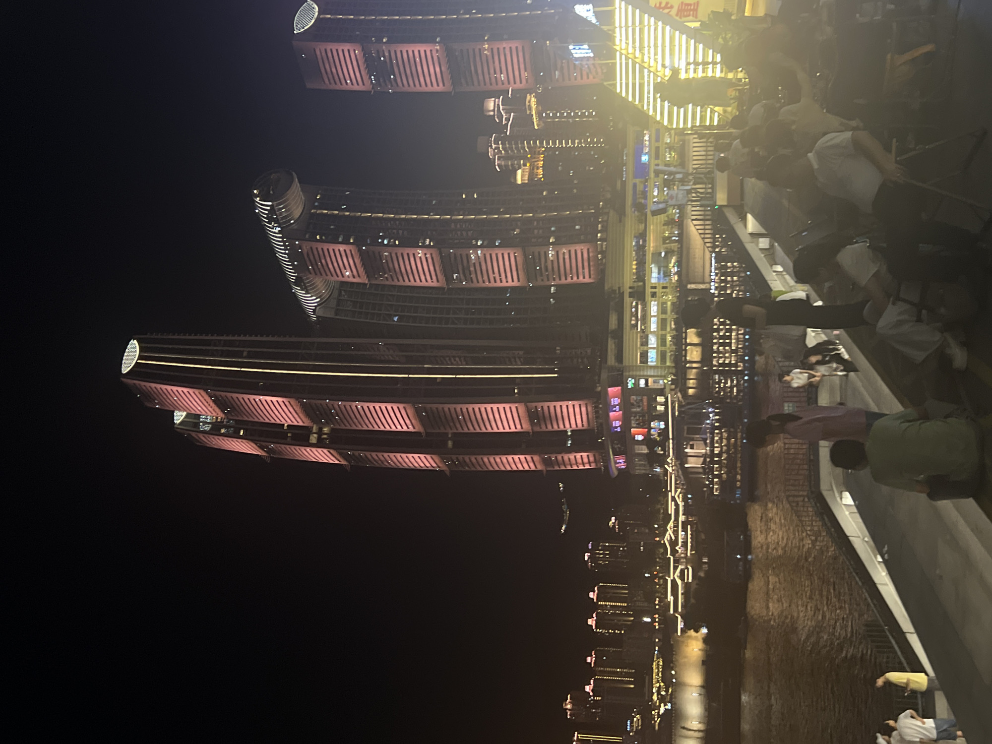 The image captures a nighttime scene along the Yangtze River in Jiangbeicheng Street, Chongqing, China. Dominating the background are several illuminated skyscrapers, prominently featuring the Raffles City Chongqing complex with its distinctive sail-shaped structures and the twin towers with pink lighting. The riverfront is lined with buildings, their lights reflecting on the water. In the foreground, a paved walkway or tiered seating area overlooks the river. Several people are present, some standing and some seated, appearing to be observing the cityscape or a performance. The atmosphere is illuminated by the city lights and the architectural lighting of the buildings.