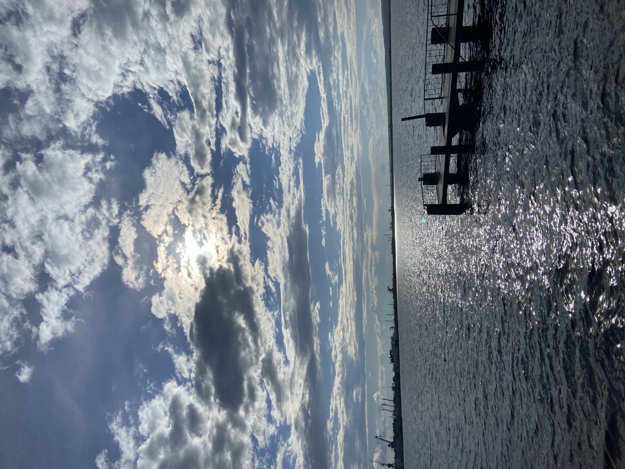 The image shows a view of the Kama River in Chaikovsky, Russia, on a partly cloudy day. The sun is peeking through the clouds, casting a shimmering reflection on the water. In the foreground, a wooden pier with metal railings extends into the river. In the distance, the silhouette of an industrial area with cranes and buildings can be seen along the riverbank. The overall scene is tranquil, with the gentle ripples of the water and the vast expanse of the sky.