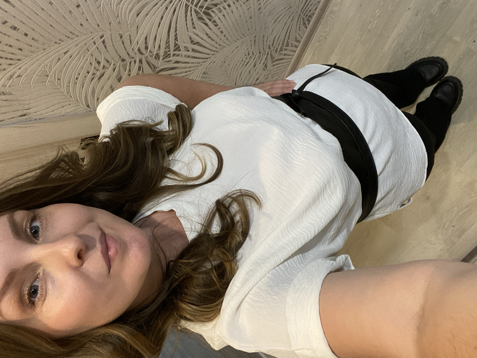 The image is a selfie of a woman lying down. She has fair skin, light eyes, and long brown hair. She is wearing a white textured shirt with a black belt or band around her midsection, black pants, and black shoes. The background is a wall with a leaf pattern and a light-colored floor. The location cannot be determined to be in Noviy, Russia from the visual data provided.