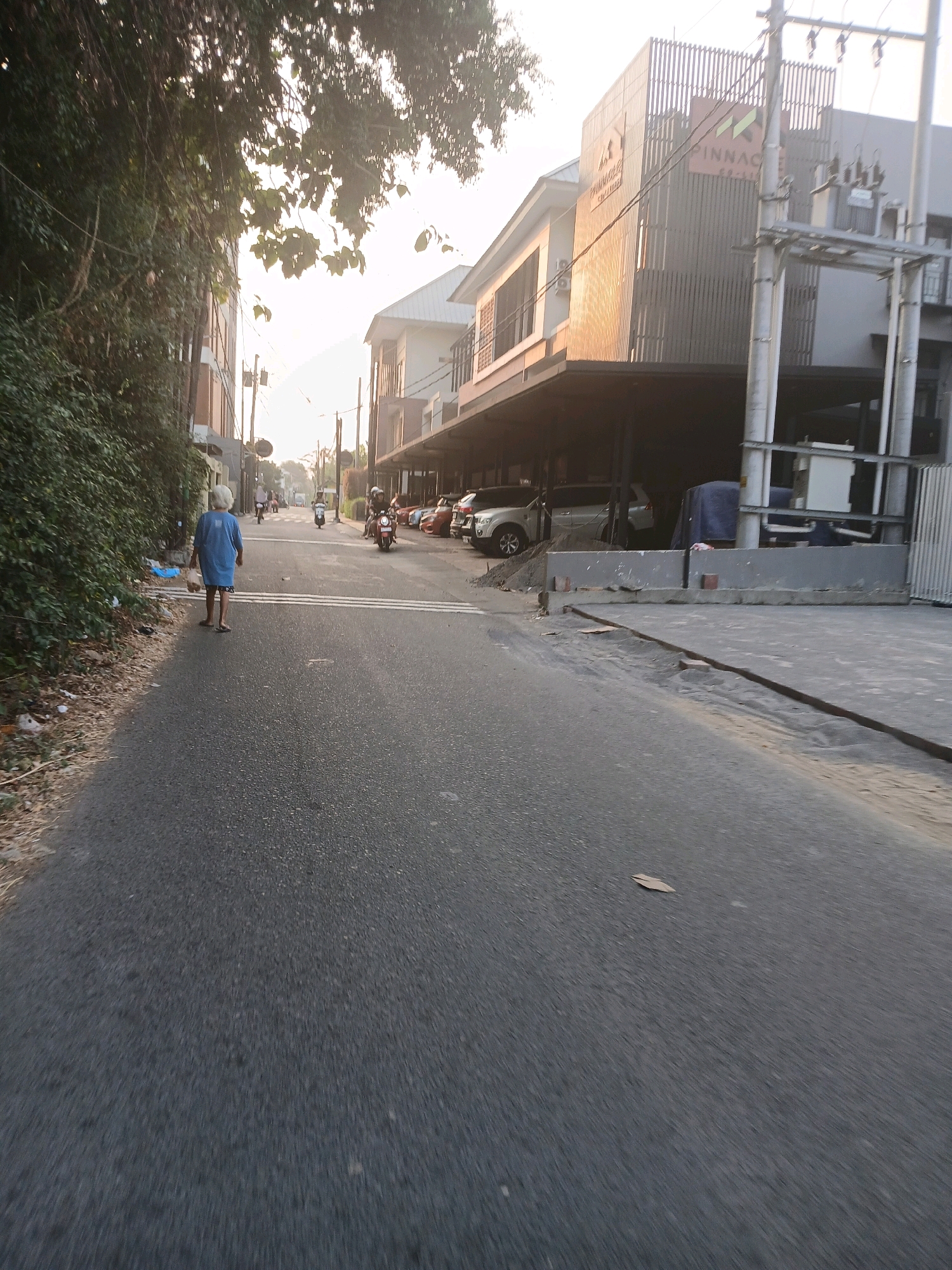 The image captures a street scene in Depok, Indonesia, under what appears to be late afternoon or early morning light. A person wearing a blue garment and shorts walks away from the camera down the center of a paved road. To the left of the road, dense green foliage covers the side of a building. Across the street, a modern, multi-story building with a facade of vertical slats and a "PINNACLE" logo is visible. Parked cars and motorbikes are present in the background, and overhead, utility poles and wires stretch across the sky. The road itself is marked with white lines, and there are some scattered leaves and debris on the side.