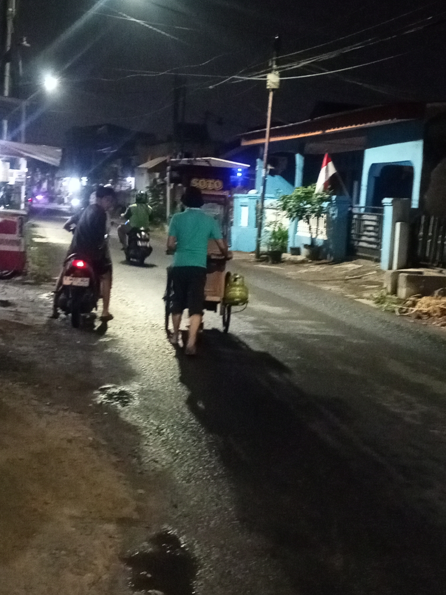 At night on a paved road in the city of Depok, Indonesia, three people are visible. In the foreground, a person wearing a dark shirt is riding a motorcycle, facing away from the camera. To the left, a red and white cart is parked. In the middle ground, a person wearing a green shirt is walking alongside a cart with a yellow propane tank attached to it. Another person is on a motorcycle behind them, also facing away from the camera. The background shows houses with blue walls and tiled roofs, a flagpole with an Indonesian flag, and streetlights illuminating the scene. The road appears wet, with some puddles.
