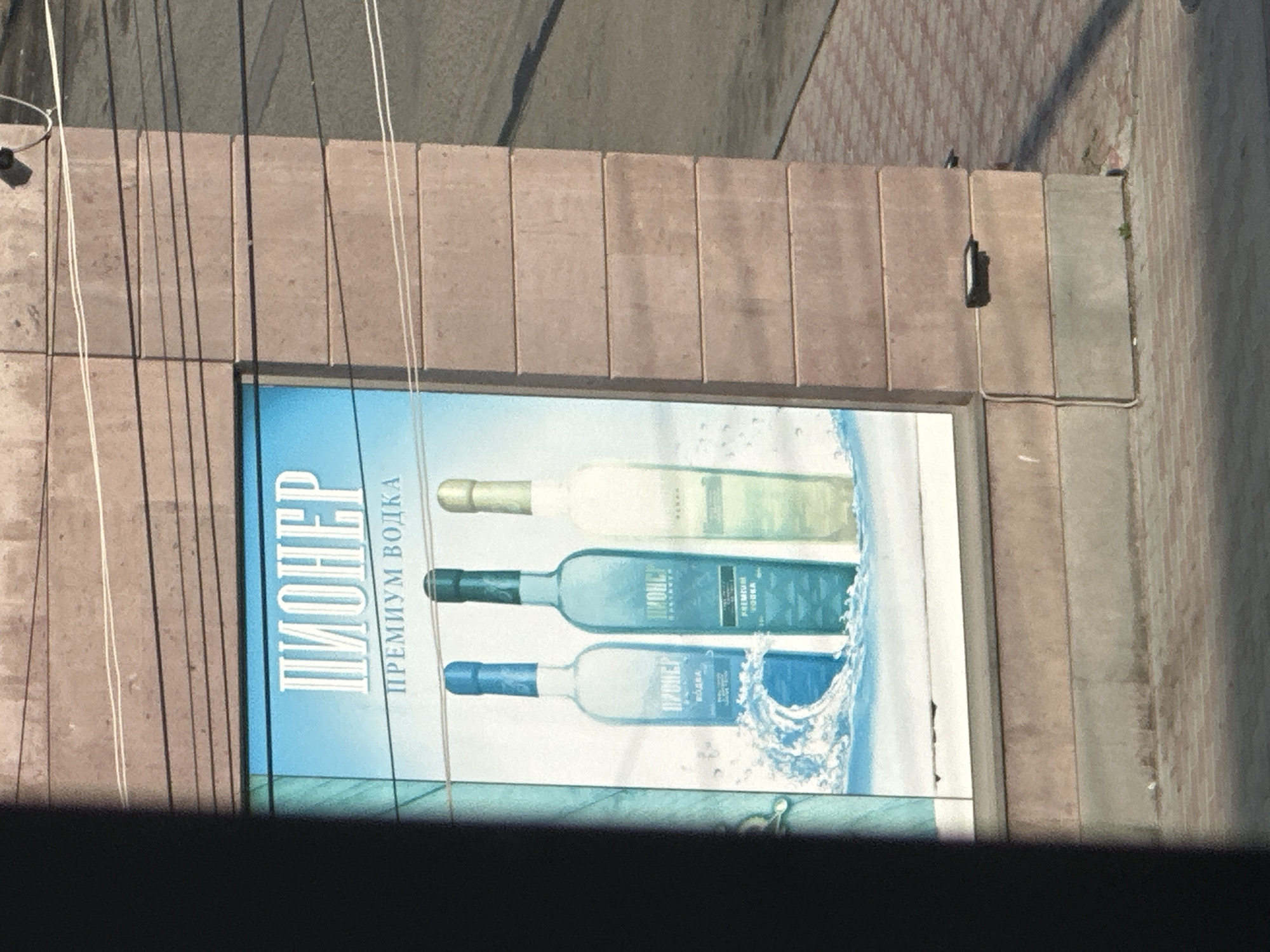 The image shows a billboard advertising "PIONER PREMIUM VODKA" with three bottles displayed. The billboard is mounted on a light brown brick building with horizontal lines. Electrical wires are visible in the upper portion of the image. The billboard itself has a light blue background with white text and a graphic of splashing water. The three vodka bottles vary in color: the left one is clear with a blue liquid, the middle one is clear with a light blue liquid, and the right one is clear with a light yellow liquid. The text on the billboard is in Cyrillic script. The lower part of the image shows a sidewalk paved with reddish-brown bricks. The overall setting appears to be an outdoor urban environment. The building and billboard are located in the city of Gyumri, Armenia, as indicated by the Cyrillic script on the advertisement.