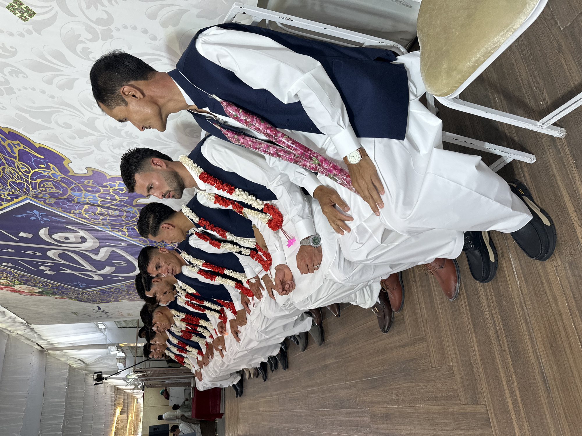 The image depicts a line of at least nine men seated in a row, presumably indoors. They are all dressed in similar attire: a white shirt and trousers with a dark blue waistcoat, and each man is wearing a garland of white and red flowers around their necks. The men appear to be in a formal setting, possibly attending or participating in an event. The men are sitting on white metal chairs with a beige padded seat. At least one of the men in the line wears a wristwatch. Each of the men are seated and are looking down.
The setting appears to be a tent or large room with a patterned ceiling in purple and gold. The floor has a wood-like finish. The location may be Quetta City Tehsil, Pakistan.