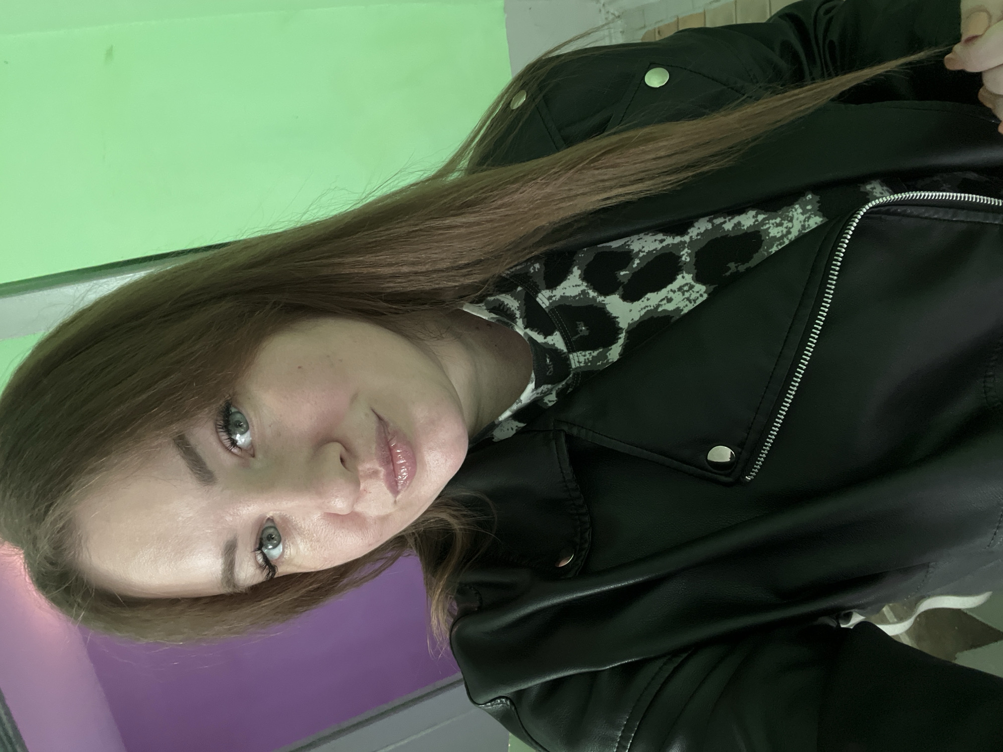 A woman with long brown hair, wearing a black leather jacket over a gray and white patterned shirt, is posing for a selfie. The background consists of a green and purple wall. The location is Chaikovsky, Russia.