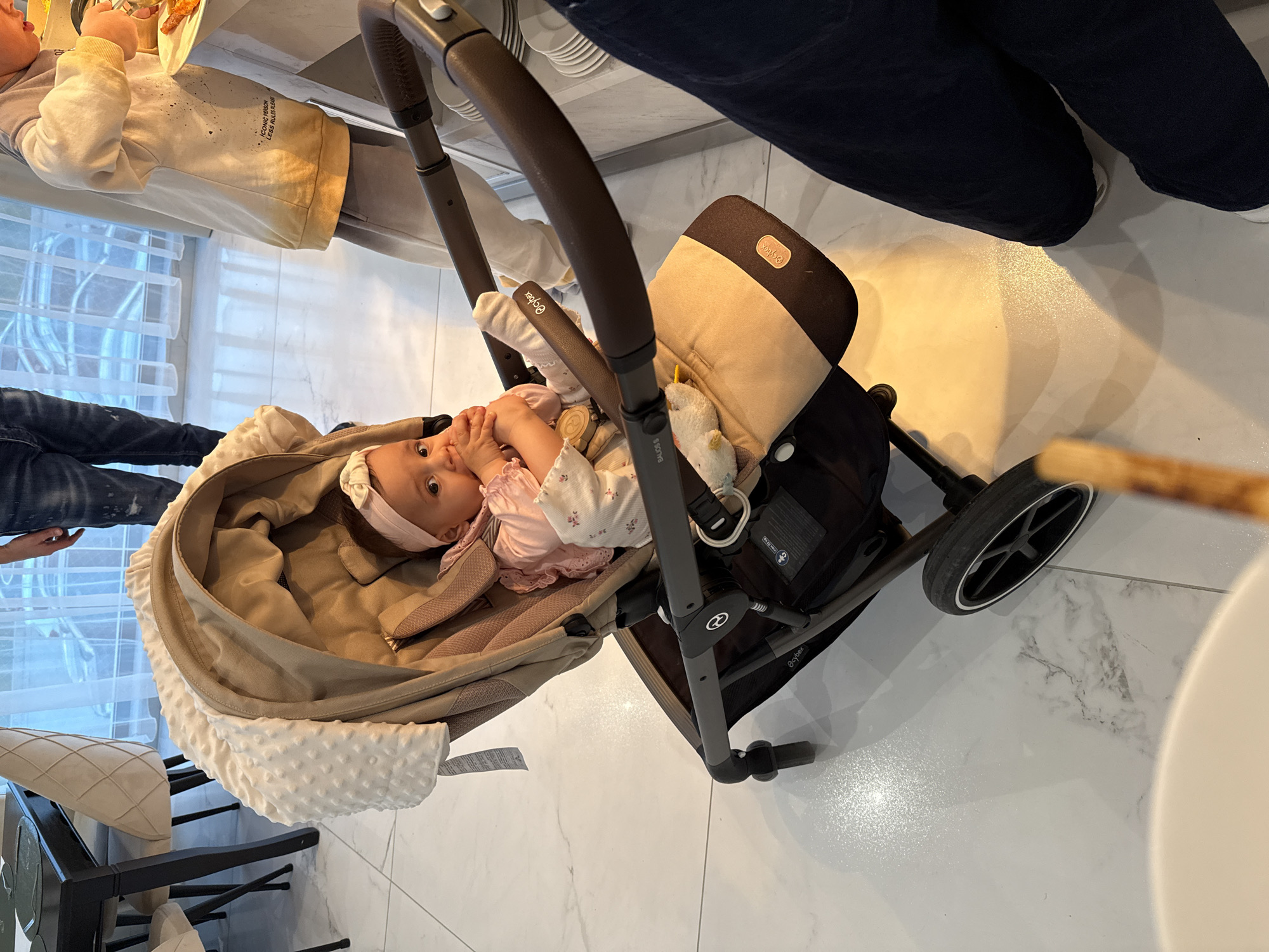 The image depicts a baby lying in a Cybex stroller, which is positioned on a white tile floor. The baby, wearing a light pink outfit and a headband, has its hands near its face. A stuffed animal is visible within the stroller. The stroller is facing away from a window with blinds, and partially obscured people can be seen in the background, including a child holding a plate and a person wearing jeans. A portion of a chair is visible in the lower left corner of the image. The location appears to be indoors, possibly in a restaurant or home dining area. The specific city or region is not identifiable from the image.