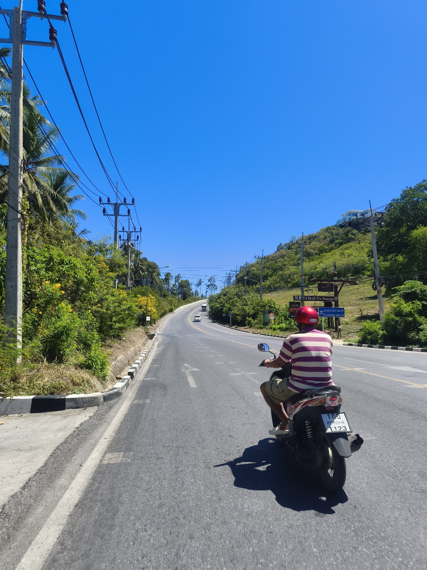 In the city of Baan Thong Po, Thailand, a person wearing a red helmet and a striped shirt is riding a motorcycle on a paved road. The motorcycle's license plate displays Thai characters and the number "1123". The road is lined with lush green vegetation and palm trees, with utility poles and power lines running alongside. In the background, a steep, tree-covered hill rises, with buildings visible at its summit. The sky is a clear, vibrant blue. Several other vehicles are visible on the road in the distance. The overall scene depicts a sunny day on a tropical roadway.