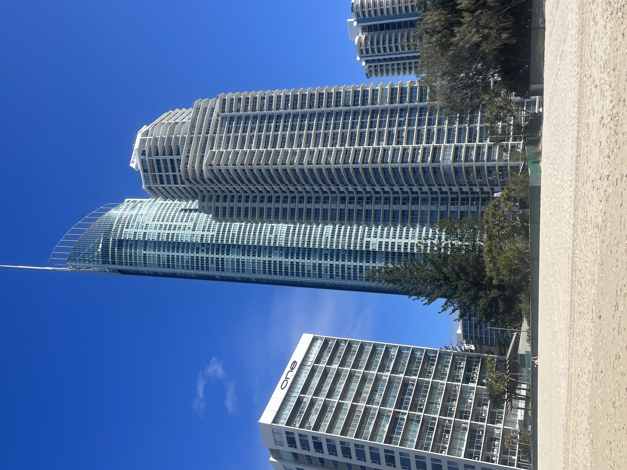 The image depicts a bright, sunny day at a beach in the Gold Coast, Australia. In the foreground, a wide expanse of light-colored sand stretches across the frame. The middle ground features lush green trees and vegetation. Behind the trees, several high-rise buildings dominate the skyline, characteristic of a coastal urban environment.
The buildings are of varying architectural styles and heights. To the left, a predominantly white building with numerous balconies displays the word "one" in large black lettering on its upper facade. Next to it, a very tall, slender skyscraper with a distinctive curved, pointed top and a facade of glass panels rises prominently. To the right of this central skyscraper, a white, multi-tiered building with many balconies is visible. Further in the background, more buildings of similar scale and style can be seen, some with curved balconies and lighter colored facades.
The sky is a clear, vibrant blue with a few wispy white clouds. A single person, appearing as a small figure, is walking on the sand in the lower left portion of the image, near the vegetation line. The overall scene conveys a sense of a modern, developed coastal city with a prominent beach area.