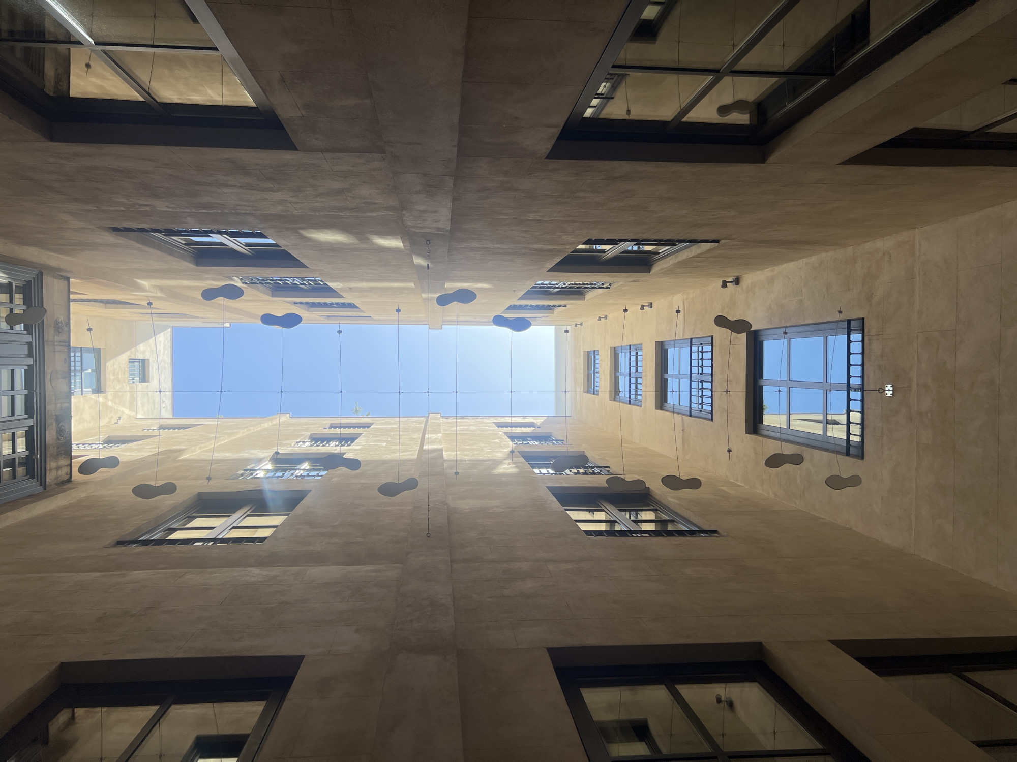 This image shows a view looking up from an interior courtyard in Barcelona, Spain. The walls of the buildings are a light, textured beige. Several windows with dark frames are visible on opposing sides of the courtyard, some reflecting the blue sky. Suspended by thin wires are dark, shoe-shaped cutouts that hang at intervals down the height of the buildings. The sky is bright blue with no clouds. The perspective emphasizes the verticality of the space, creating a sense of looking up towards the sky.