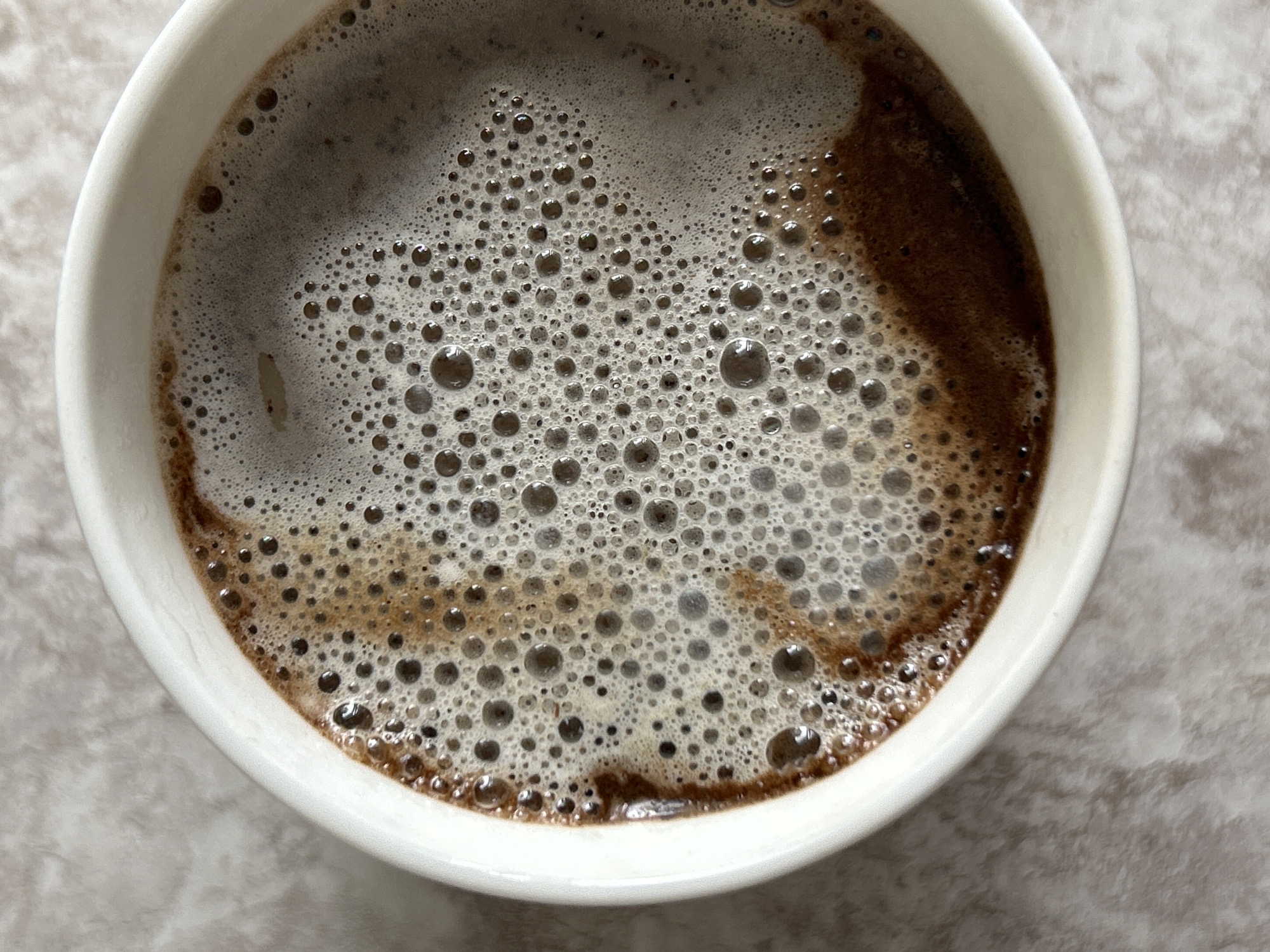 A close-up, overhead shot focuses on the foamy surface of coffee in a white mug. The coffee has a rich brown hue and is covered in a layer of small, irregular bubbles, some darker than others. The mug itself is a simple white ceramic, with its rim partially visible in the foreground and on the left. The background appears to be a textured, light-colored surface, possibly a countertop or placemat. The image was taken in Chaikovsky, Russia.
