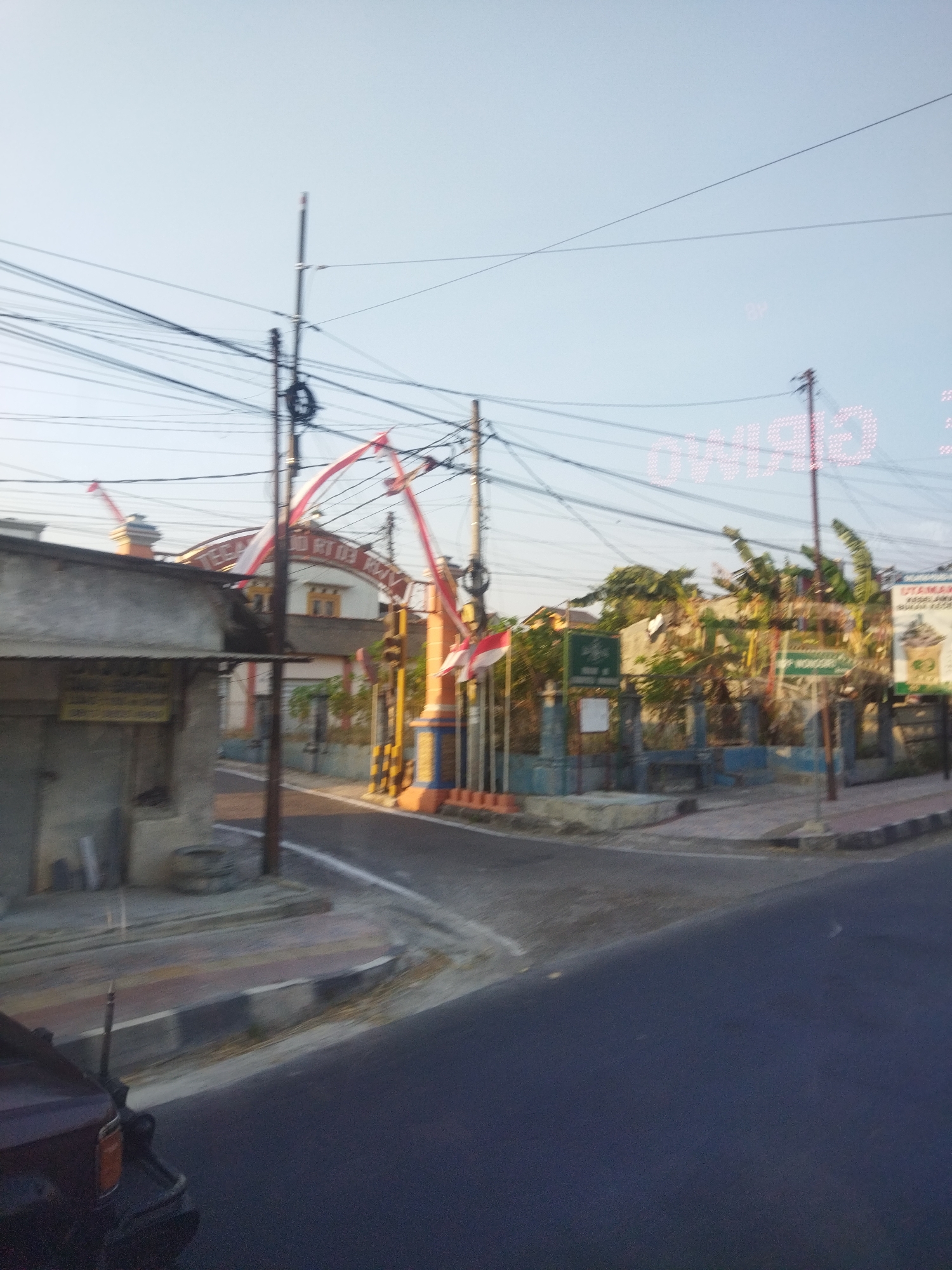 The image depicts a street scene in Wonokarto, Indonesia, likely in the morning or late afternoon given the lighting. The foreground shows a dark asphalt road, with a portion of a dark-colored vehicle visible on the left. The road curves slightly to the right, leading towards a built-up area.
The midground features a street with a raised sidewalk and a pathway. On the left side of the street, a small, weathered building with a concrete facade is visible. A sign with illegible text is attached to its wall. Further along the street, on the right side, is an entrance to a compound or property. This entrance is marked by a large, arched gateway adorned with red and white Indonesian flags. Behind the gateway, a building with a white and orange facade is partially visible, along with lush green trees and vegetation. Several poles with a multitude of electrical wires stretch across the sky, creating a dense network overhead. A prominent, illuminated sign with pink lettering, partially obscured and reversed due to the viewing angle, is positioned in the upper right quadrant of the image. Below this sign, another, more distant sign with what appears to be an image of a drink cup is mounted on a fence. The overall impression is that of a street in a suburban or semi-urban area in Indonesia, with a mix of residential, commercial, and public spaces.
