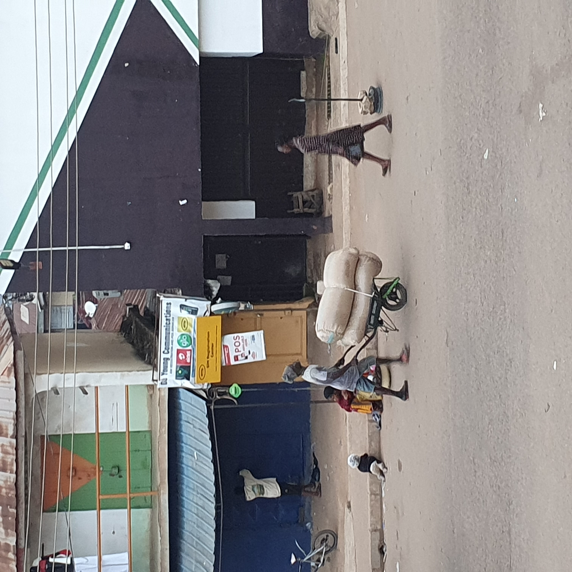 A medium shot shows a street scene in a city. In the foreground, a man with a head wrap and light-colored shirt and blue shorts pushes a two-wheeled cart loaded with sacks. To his right, a woman wearing a striped dress walks across the street. In the background, a baby is sitting on the side of the street, and a young child in a red and yellow outfit is sitting next to the baby. Further back, a person in a light-colored shirt is painting a blue wall. A bicycle is parked on the left side of the image. A building with a dark brown facade and a white section with green stripes runs along the top right of the image. A smaller building with a blue awning and a green door is on the left. A sign for "Young Communications" is visible on the right side of the image.