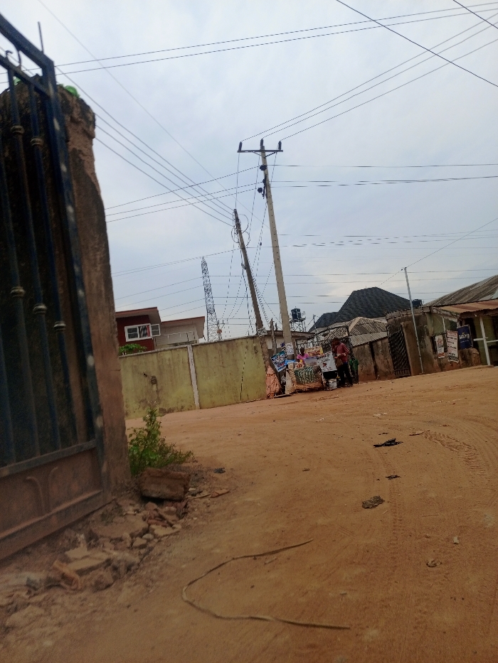The image depicts an unpaved, reddish-brown dirt street in Magboro, Nigeria, under an overcast sky. In the foreground, a dark vertical metal gate component is visible on the left, secured to a rough concrete pillar with scattered concrete debris and sparse green vegetation at its base. A dark, thin linear object, likely a wire or cord, lies on the dusty ground, which exhibits tire tracks.
In the midground, on the right side of the street, an informal roadside kiosk is present. It features a light-colored surface, likely a table, displaying various packaged goods and multiple posters or advertisements. Two individuals are observable near this kiosk: one person wearing a light-colored top (possibly pink/purple) and another in a red top. Their positioning suggests interaction with the kiosk, either as a vendor or customer.
The background shows an urban environment. A pale building with a red-framed window is partially visible behind a low, yellow-green concrete wall. Multiple utility poles, one concrete and one leaning, support numerous overhead electrical transmission lines that crisscross the frame. A steel lattice transmission tower is discernible in the distant background. Various structures with different roof types, including dark shingles and corrugated metal, line the street. A water storage tank on a stand is visible above a building roofline. Additional advertisements are affixed to background walls.