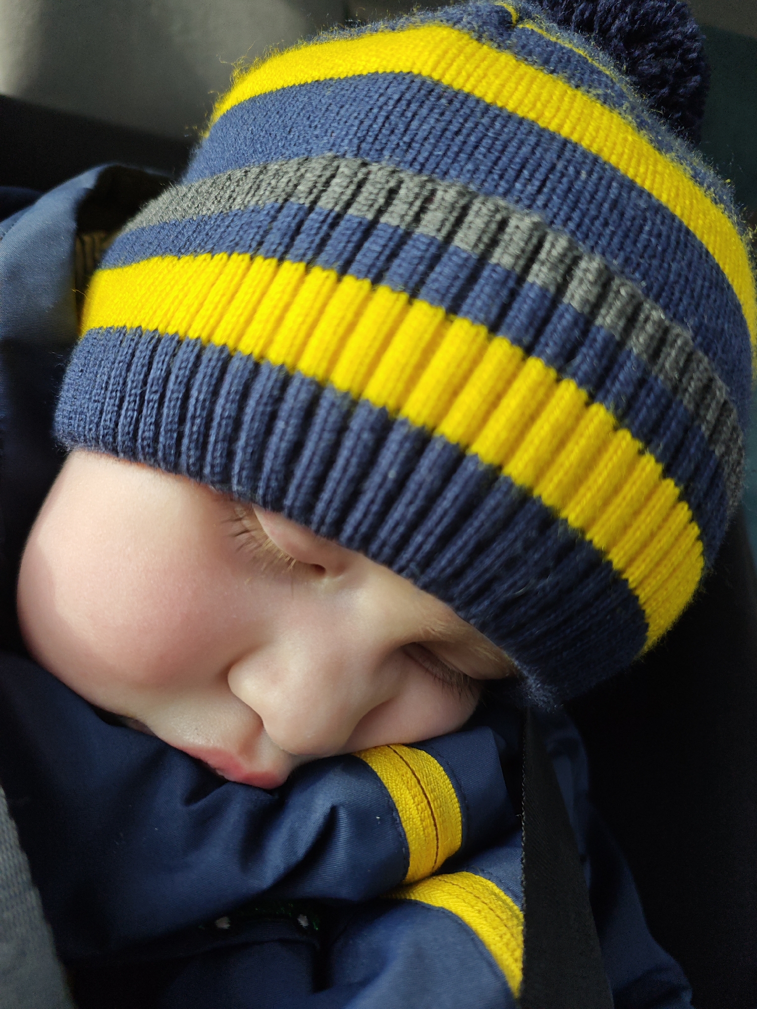 An infant or young child is depicted sleeping in a close-up, angled portrait. The child wears a knitted hat with horizontal stripes in navy blue, bright yellow, and grey, topped with a dark blue pompom. A navy blue outerwear garment, possibly a jacket or snowsuit, with a visible yellow stripe, covers the child's body. The child's head is tilted forward, resting on the blue garment. The visible right side of the child's face shows closed eyes with light eyelashes, prominent rosy cheeks, and slightly parted lips. A distinct red mark is present on the upper lip area. A dark, possibly black, strap consistent with a safety harness is visible on the right side, suggesting the child is secured in a carrier or seat. The background is dark and out of focus. The scene is located in the city of Chaikovsky, Russia.