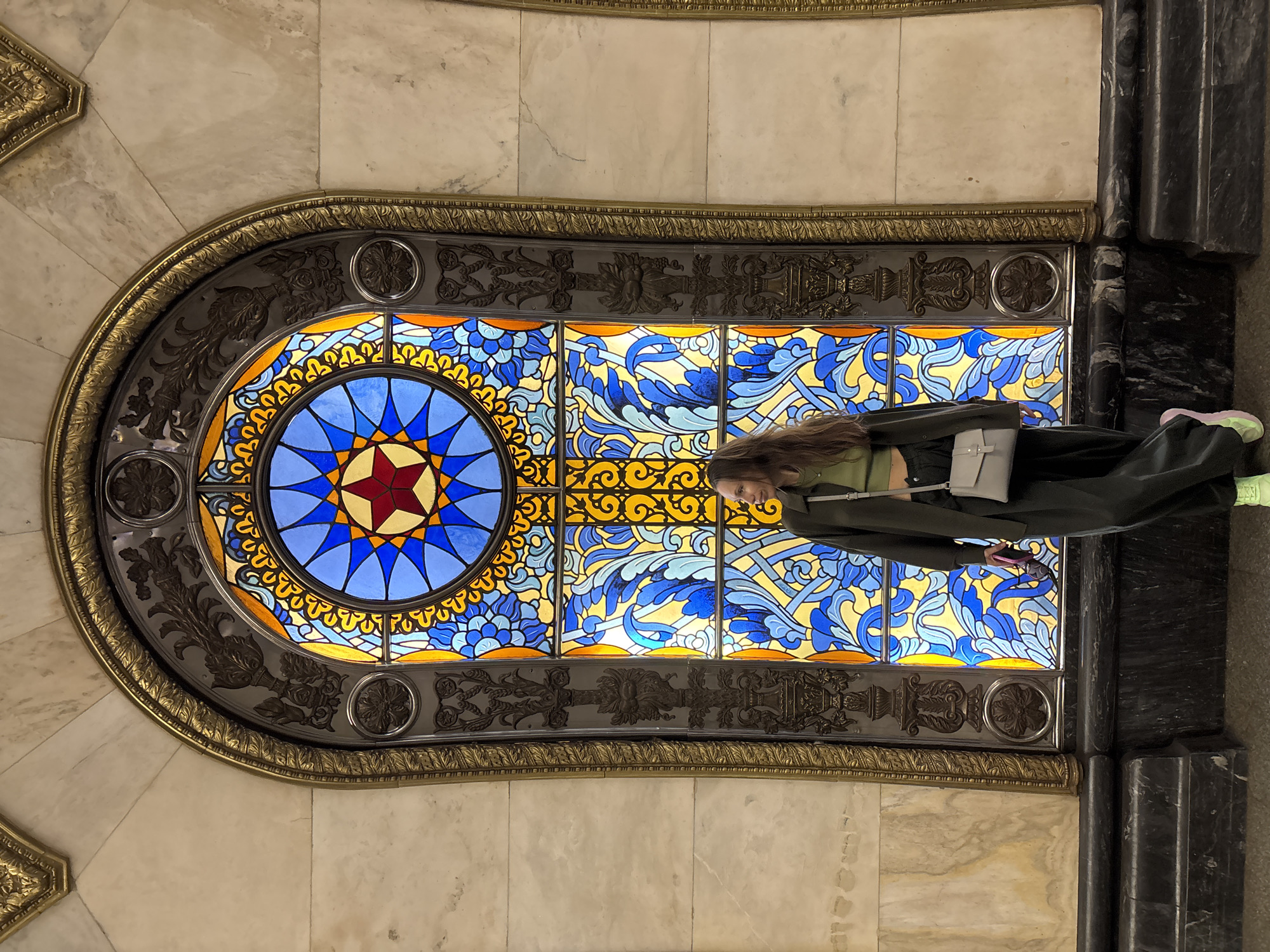 The image shows a large, ornate stained-glass window in what appears to be a Moscow Metro station. The window is horizontally oriented and features a complex design with a central circular motif of blue triangles radiating from a red star. The remainder of the window is filled with swirling blue and yellow patterns resembling foliage or waves. The window frame is made of dark metal adorned with detailed floral and geometric patterns, and edged with gold trim. The surrounding walls are clad in marble. A person is standing in front of the window. They are wearing a green shirt, a dark jacket and dark pants. They have a light-colored shoulder bag. Their face is partially visible. A hint of lime green shoes is visible near the edge of the frame. A dark marble pillar is to the right of the window.