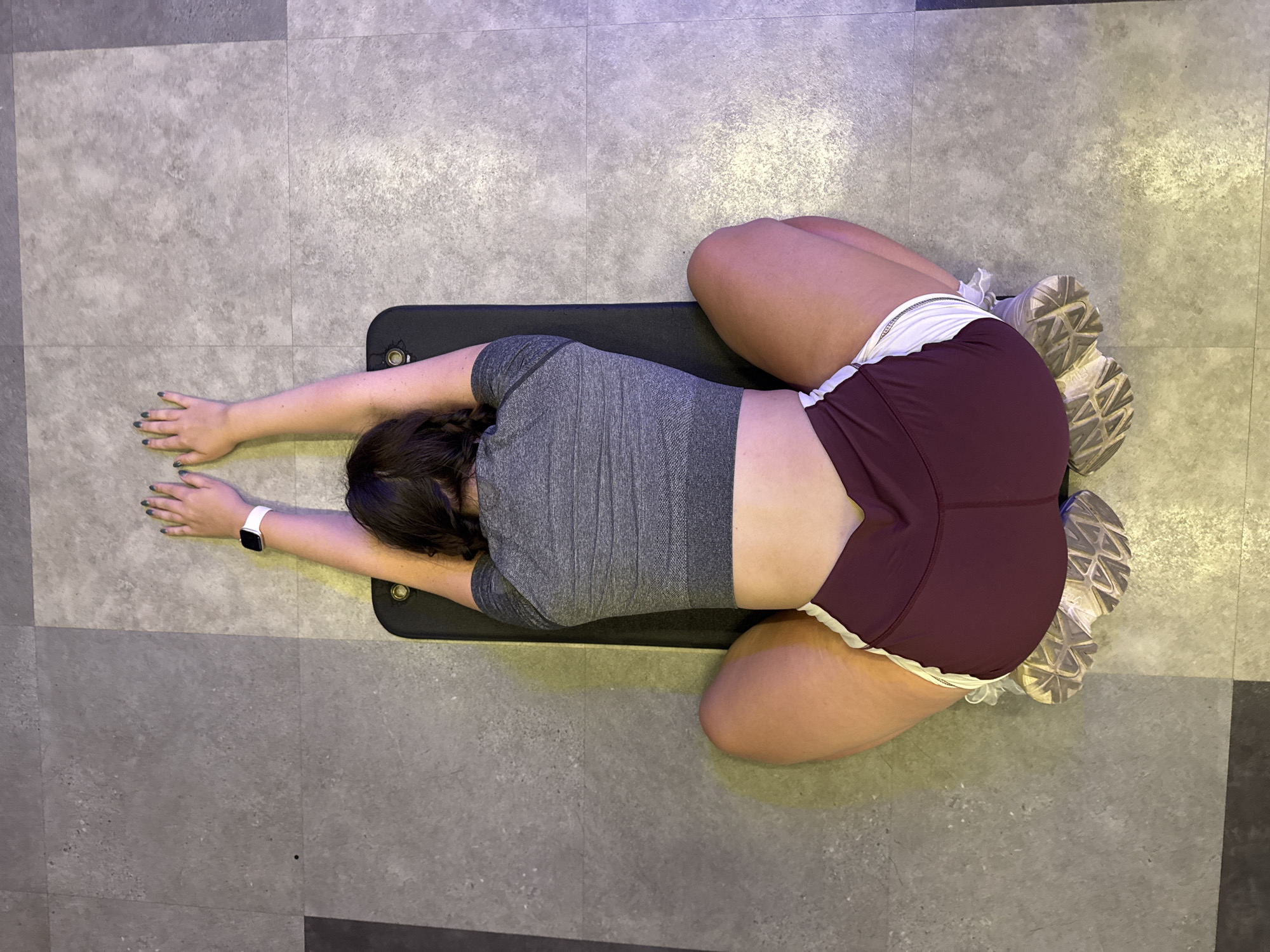 A person with dark hair is lying on their back on a black exercise mat, which is placed on a gray tiled floor. The person is wearing a gray, cropped, short-sleeved shirt and maroon shorts with a white trim. Their arms are extended overhead with their palms facing upwards. Their legs are bent at the knees, with the soles of their feet resting on the floor. The scene is dimly lit with a purple light emanating from the right side of the image. The location is identified as Nong Pa Khrang, Thailand.