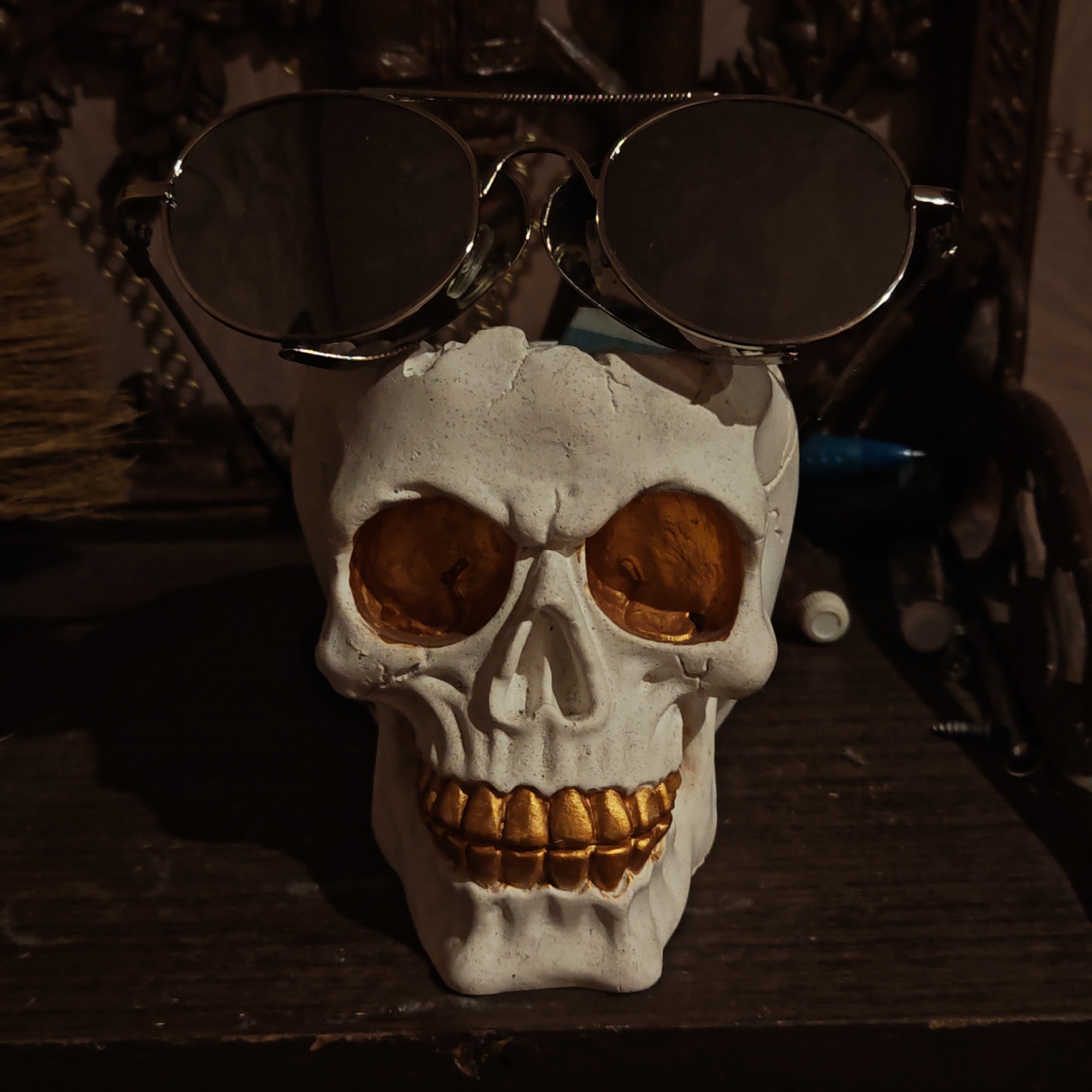 A white skull with gold teeth and eye sockets rests on a dark, wooden surface. A pair of sunglasses with gold frames are perched on the skull's forehead. In the background, there is ornate detailing. There is no indication of location.