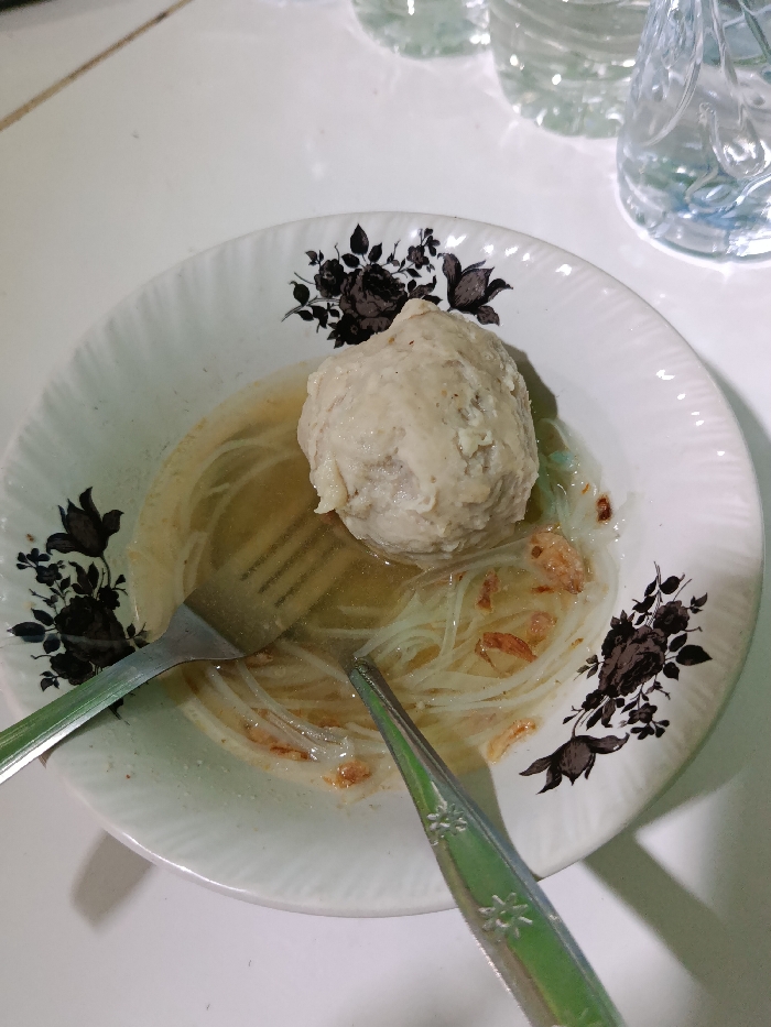 A white ceramic bowl with a black floral pattern, containing a large meatball in a clear broth with thin white noodles and fried onions. The bowl is on a white surface. Two forks lay on the side. The location, based on the coordinates, is a residential area in the city of Kecamatan Arjasa, Indonesia. The image suggests a meal in progress. The bowl is positioned near the center of the image. The meatball is visible, lying in the broth. The forks appear to have been used. The image implies a simple, informal meal setting.