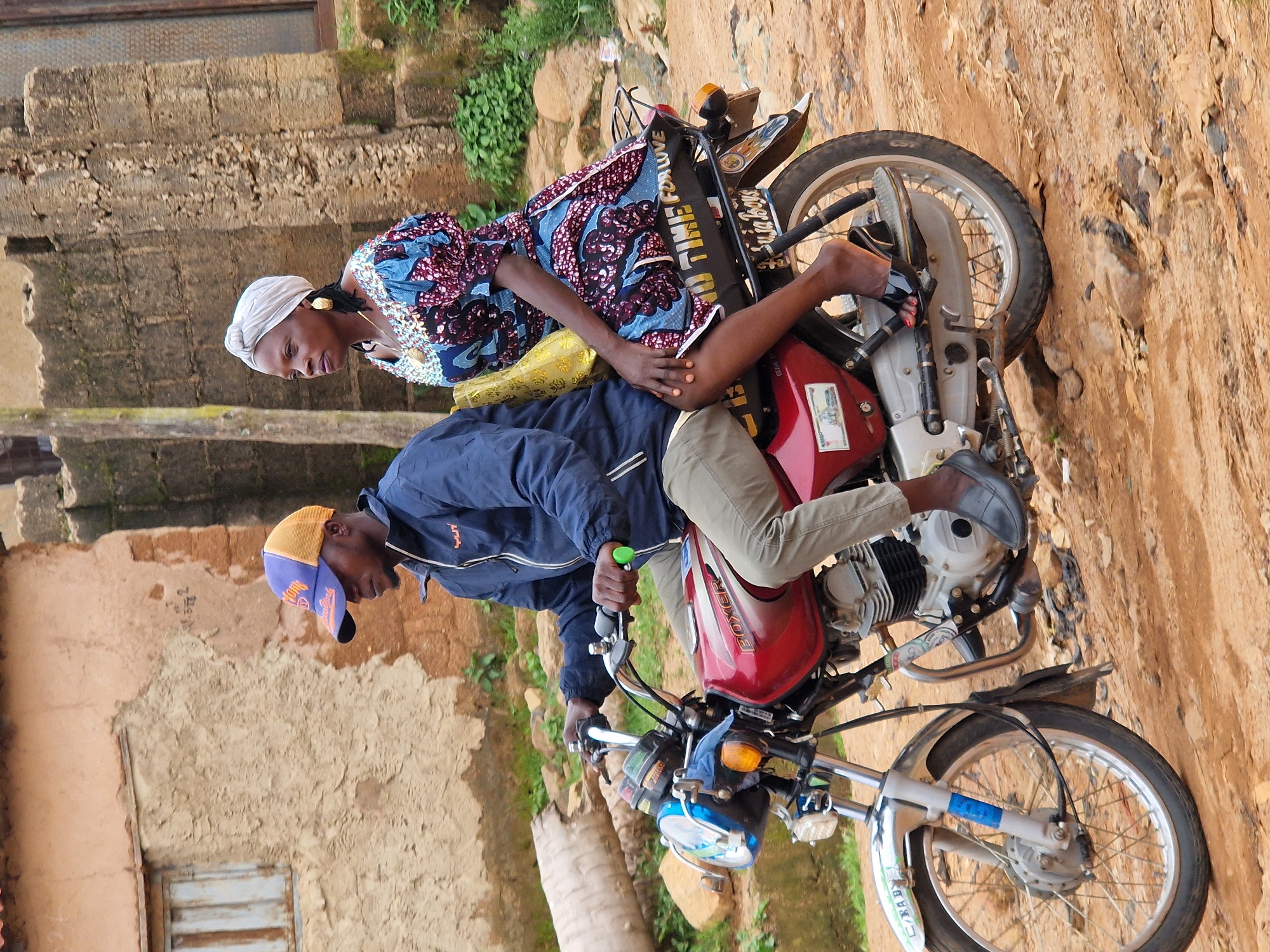 Two people are riding a red motorcycle on a dirt road in front of a weathered building. The driver, a man wearing a blue jacket and a blue and orange baseball cap, is looking forward. The passenger, a woman with a white headwrap and a colorful patterned dress, is looking to her left. The motorcycle has "BOXER" written on its side. The building behind them has a rough, textured facade and a small window. Green foliage is visible in the background. The scene appears to be in a rural or semi-urban setting.