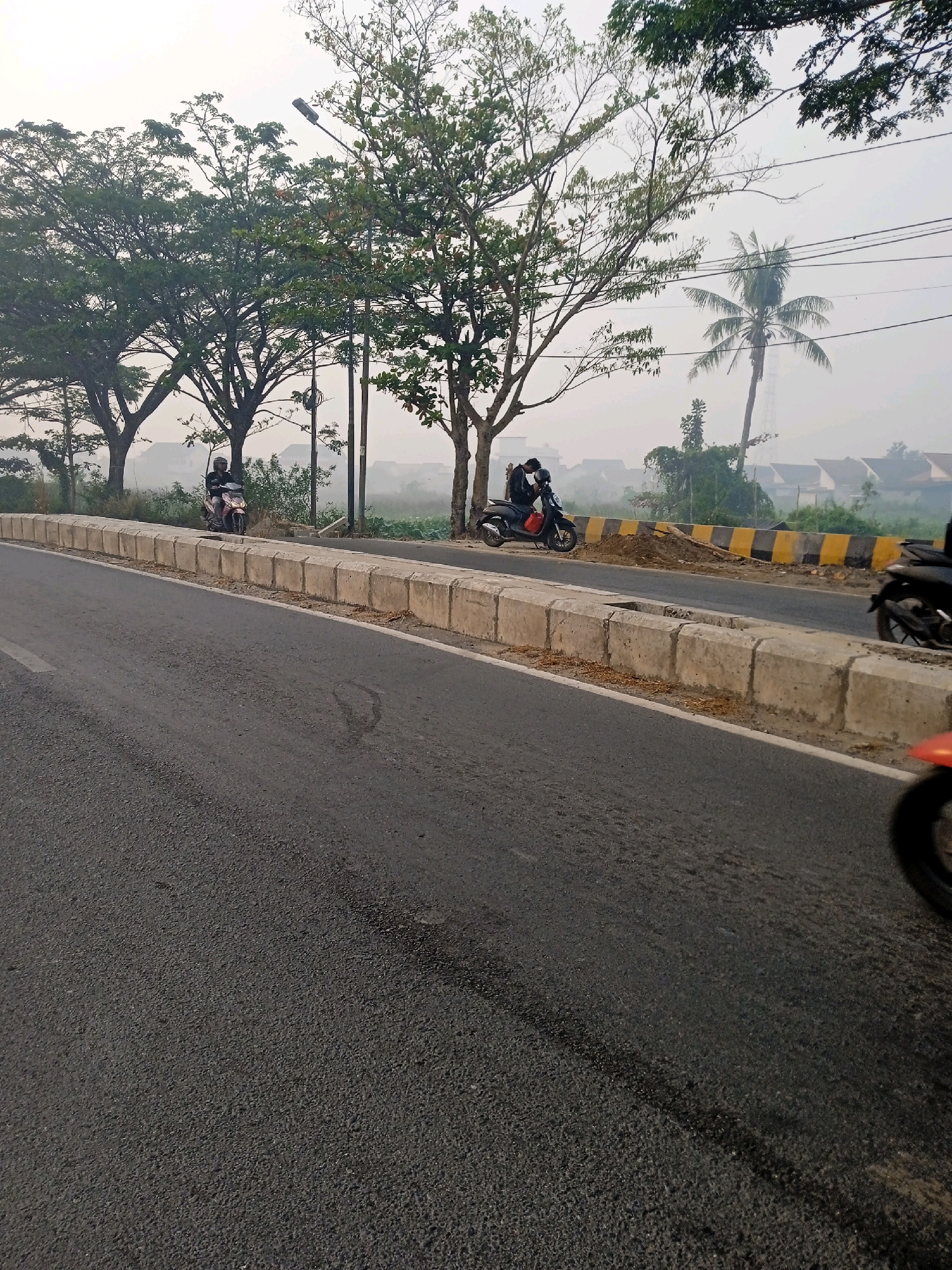 A view of a street in Banjarmasin, Indonesia, on a hazy morning, featuring a road with concrete dividers. Two motorcycles with riders are visible on the side of the road, one with a person wearing a helmet and a black jacket on a red and white motorcycle, and another with two people on a black scooter. Beyond the road, there are trees, a palm tree, a utility pole, and residential buildings in the distance. Yellow and black striped barriers are also present on the side of the road. The weather appears to be overcast or foggy.