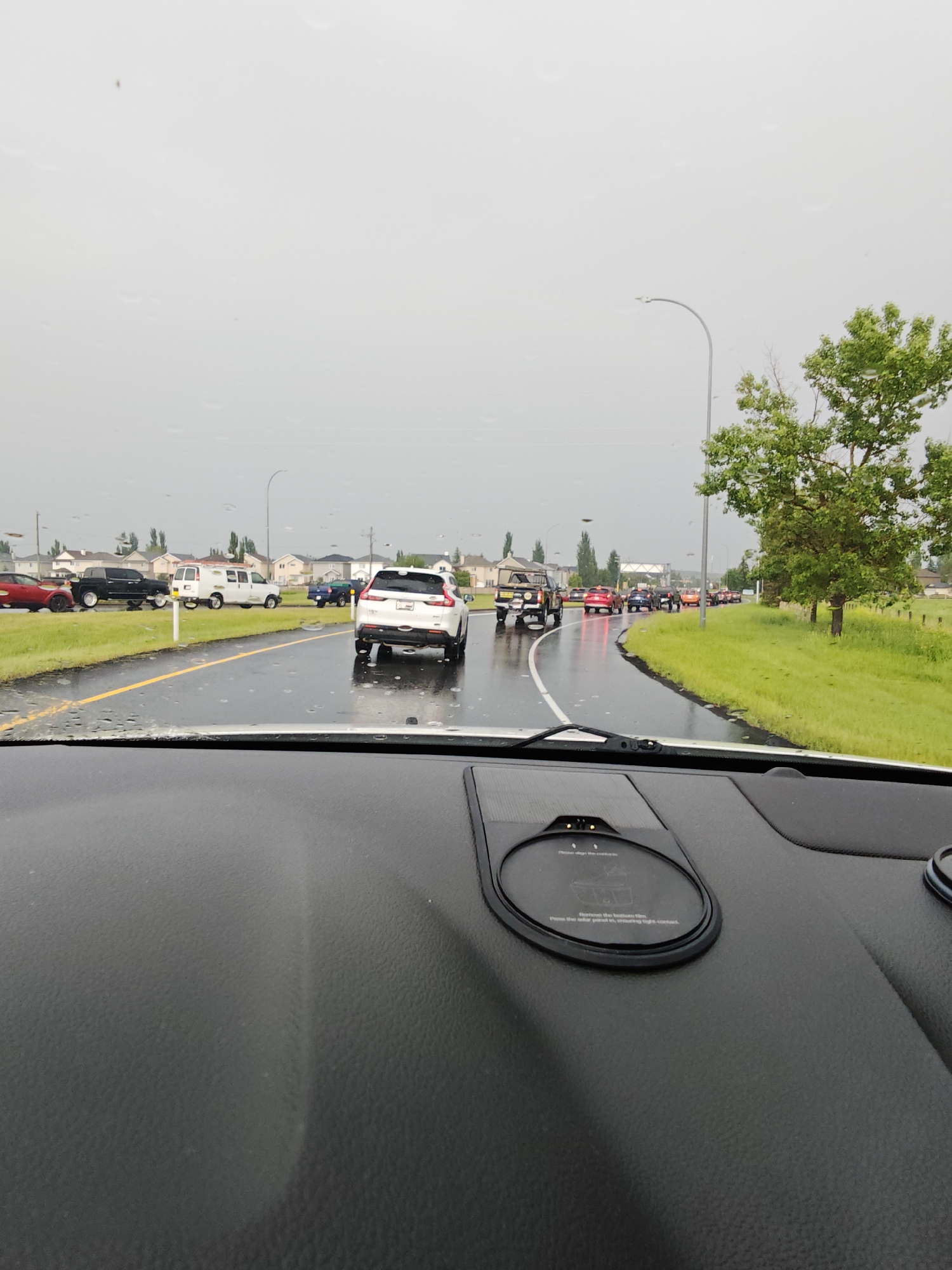 The image captures a traffic scene in Calgary, Canada, on a rainy day. The view is from the interior of a vehicle, looking out through a wet windshield. A line of vehicles is visible on a wet asphalt road with yellow lane markings. Cars of various colors, including white, black, red, and blue, are stopped or moving slowly. A white SUV is in the foreground on the right, and a white van is further ahead on the left. In the background, residential houses with light-colored siding and dark roofs line the street, characteristic of many suburban neighborhoods in Calgary. Lush green grass and trees flank the road, suggesting a summer or late spring season. The sky is overcast and grey, consistent with rainy weather. A street lamp is visible on the right side of the road. The dashboard of the vehicle in the foreground is partially visible, showing a dark, textured surface and what appears to be a wireless charging pad.