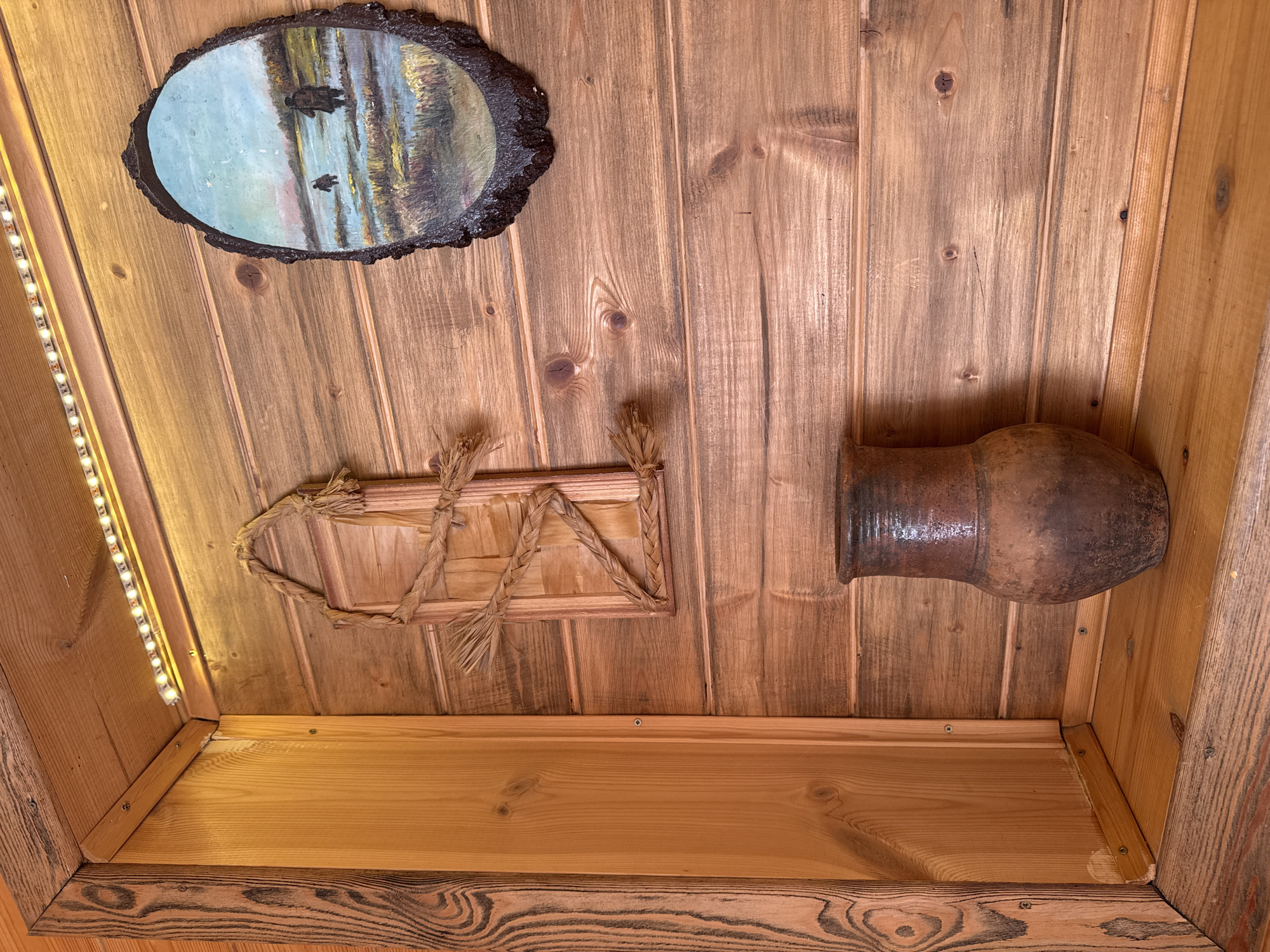 A low-angle shot shows the interior of a wood-paneled structure. In the upper left, there's a framed painting on a slab of wood. Below it, LED strip lights run vertically. To the right of the painting is a framed straw decoration. In the upper right, there is a dark clay pot attached to the paneling. At the bottom of the image, a shelf constructed of light-colored wood is visible. The background consists of brown-toned, aged wood paneling with visible knots. There is no mention of the location in the city of Ketoshi, Russia.