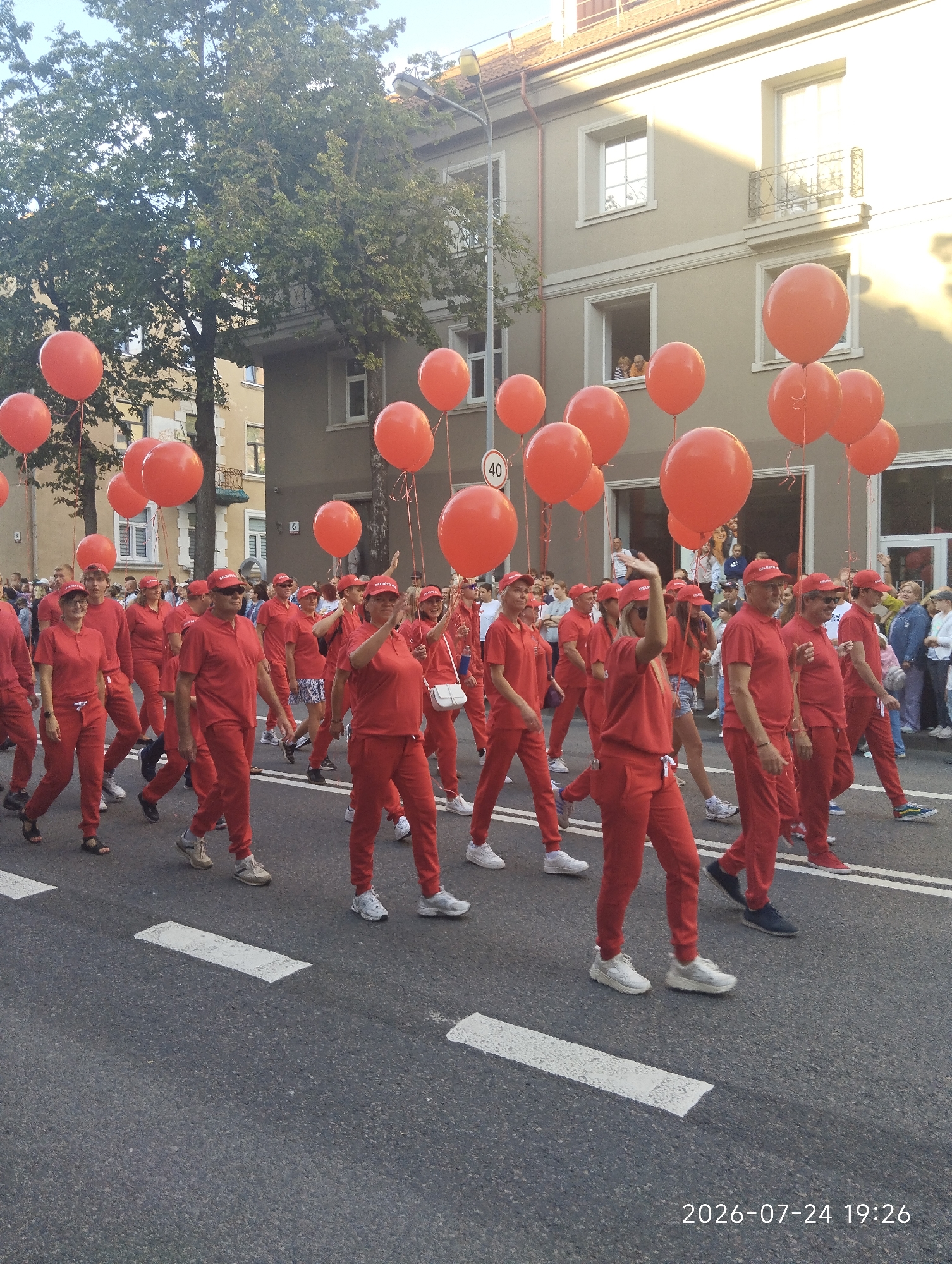 A group of people in matching red outfits and red hats are walking in a parade. Many of them are holding large orange balloons. The parade appears to be taking place on a city street, with buildings and trees in the background. The sun is shining, casting long shadows. A sign with the number "40" is visible on the left side of the image. The date stamp in the bottom right corner indicates the image was taken on July 24, 2026, at 19:26. The specific location in the city of Klaipėda, Lithuania, is not explicitly stated but can be inferred to be a public street likely hosting a celebratory event.