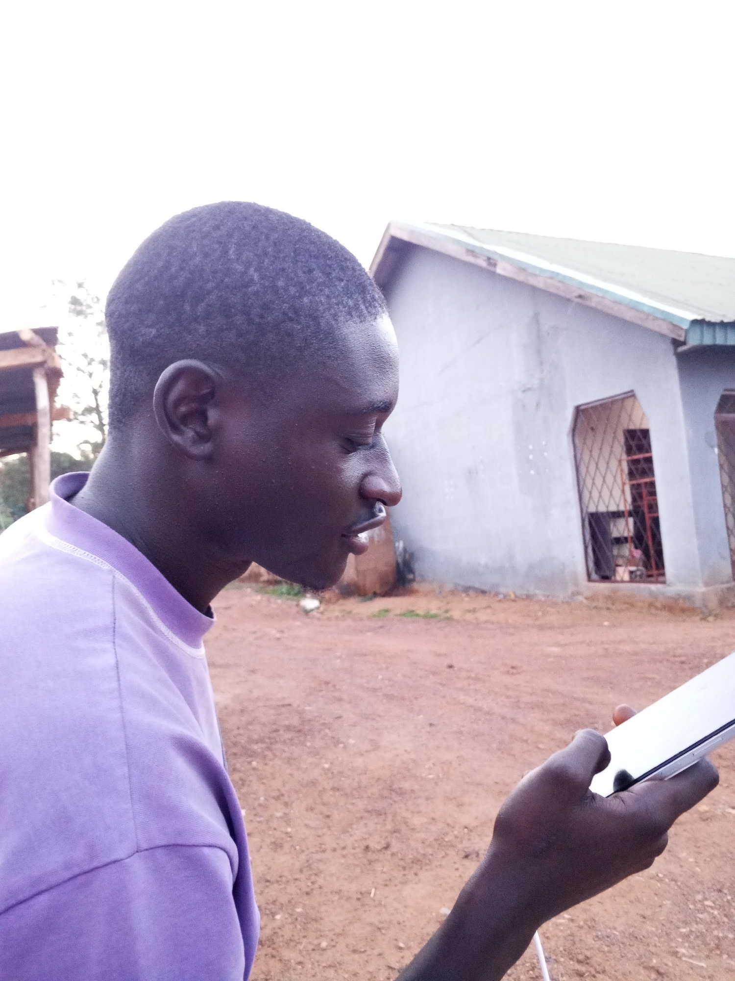 A dark-skinned man is holding a smartphone, viewed in profile, outdoors. He is wearing a purple t-shirt. In the background is a gray building with a metal-barred window and a roof with green trim. The ground appears to be red dirt. The location is Bukuru, Nigeria.