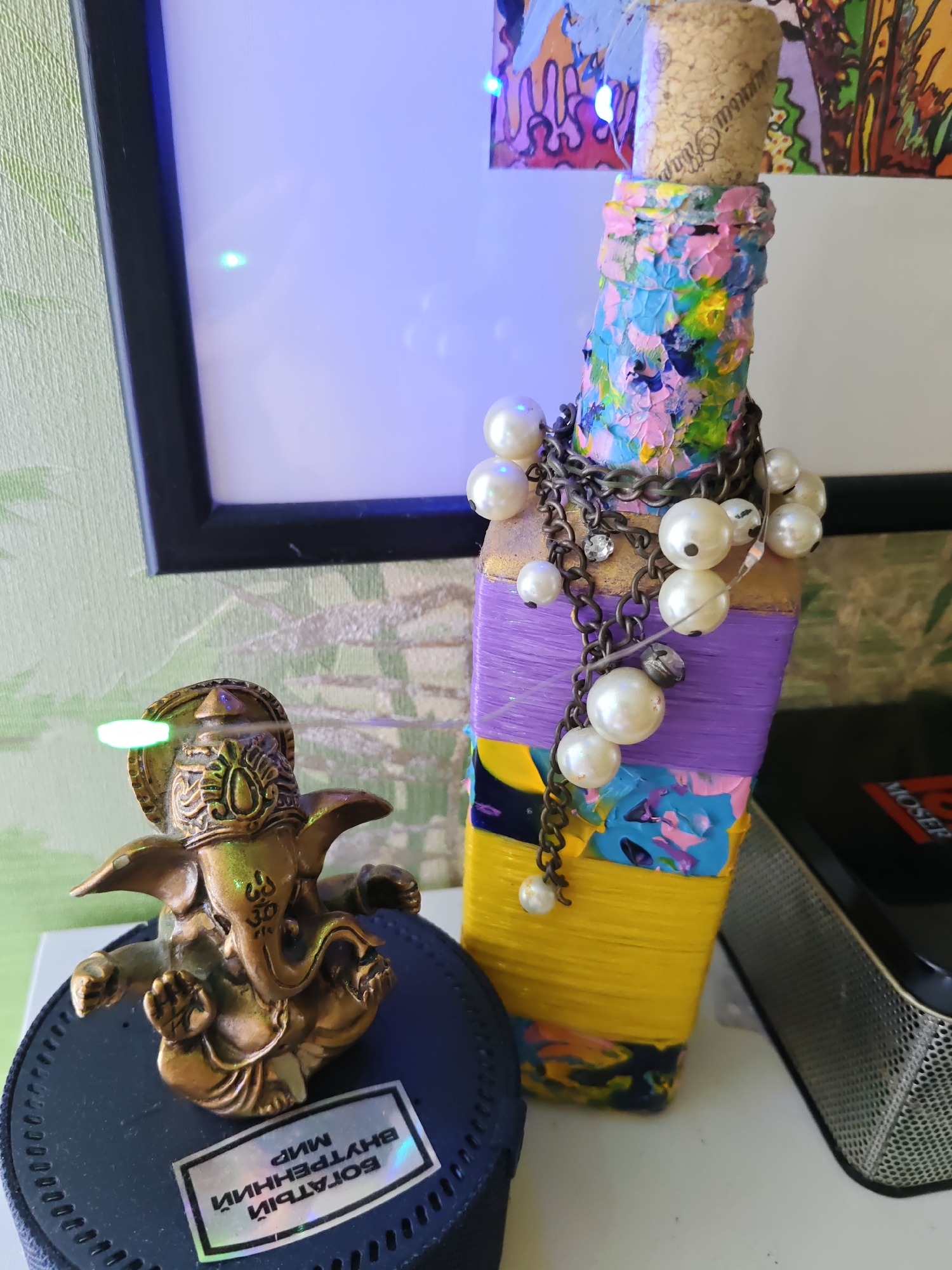The image depicts a tabletop scene featuring a golden Ganesha figurine and a decorative bottle. The Ganesha figurine is positioned on a dark circular base with a holographic sticker that has Cyrillic text. Adjacent to it is a bottle wrapped in yellow and purple yarn, with colorful paint accents and adorned with pearl beads and chains. The background shows a framed picture and a textured wall. The scene appears to be an indoor setting, possibly a room in Rostov-on-Don, Russia, given the presence of Cyrillic text on the sticker, though the specific location within the city cannot be determined from the image alone. There are no people or discernible actions occurring in the image. The lighting suggests ambient room illumination, with some artificial lights reflecting off the subjects.