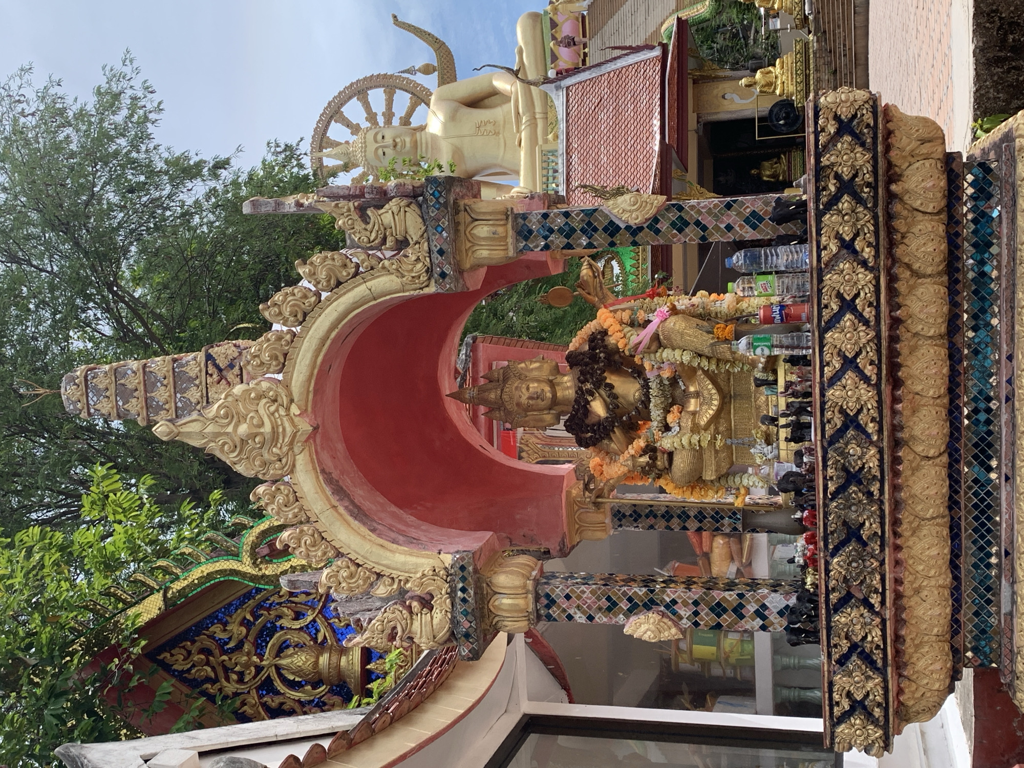 In the city of Ko Samui District, Thailand, a golden statue of a multi-faced deity, adorned with garlands of flowers, sits prominently on an ornate, gilded altar. The altar itself is richly decorated with intricate patterns and colorful mosaic tiles. Behind the deity, a large golden Buddha statue is visible, seated majestically on a tiled roof structure. The surrounding environment features lush green trees and a glimpse of a cloudy sky, suggesting an outdoor setting at a temple or shrine. The scene is bathed in daylight.