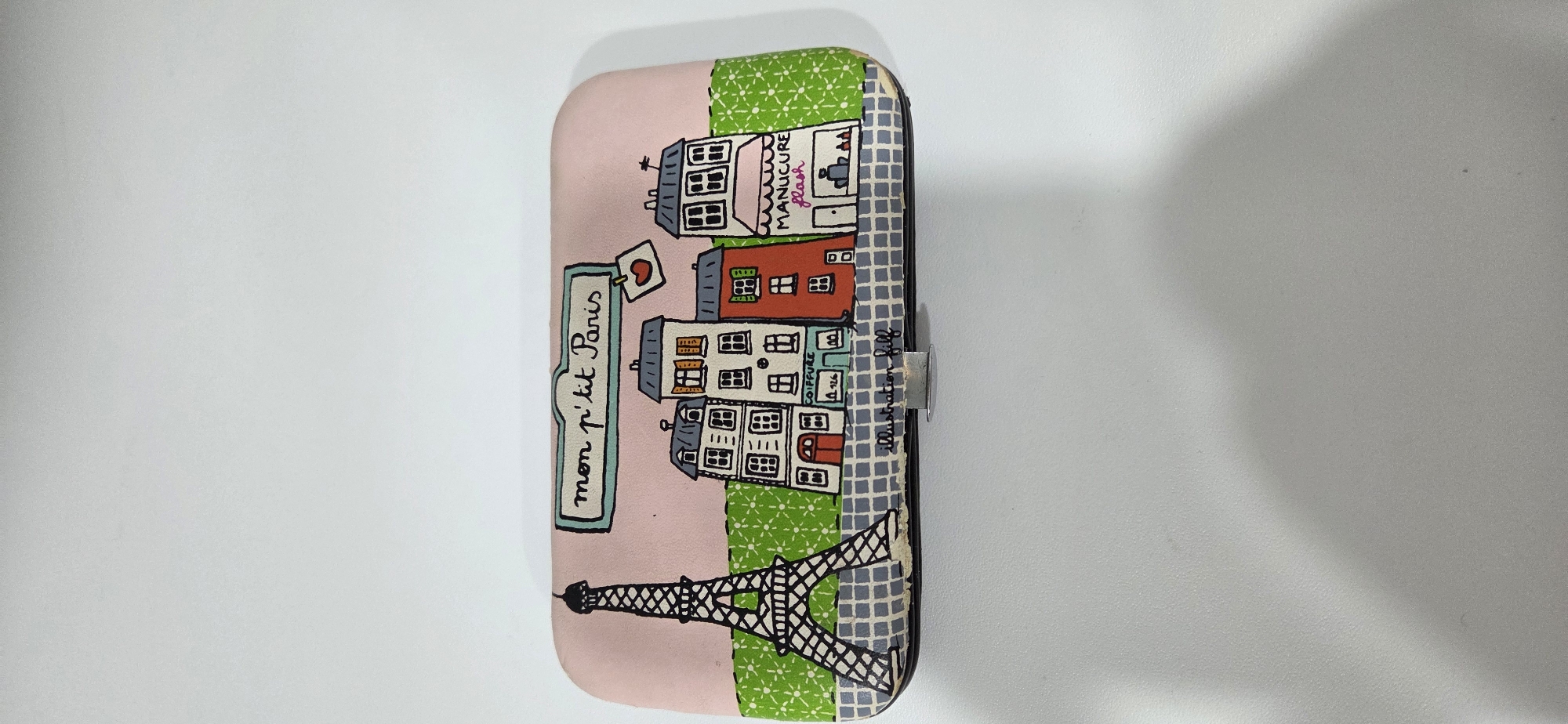 A rectangular object, likely a case or box, with rounded corners, is depicted resting on a plain white surface. The object's top surface features a whimsical, hand-drawn style illustration of a Parisian street scene. The background of the illustration is pink, transitioning to a green patterned strip and a gray and white checkered base. On the left, a black-outlined Eiffel Tower is visible. To its right, a series of cartoon-style buildings are drawn, including one labeled "COIFFURE" with "A 126" below, and another with a pink awning labeled "MANUCURE flash". Above the buildings, a banner reads "mon p'tit Paris" in black script, accompanied by a small red heart. The signature "illustration Jill" is visible on the lower edge of the illustrated surface. A metallic clasp mechanism is positioned centrally on the front bottom edge of the object. The object is situated in the city of Lipetsk, Russia.