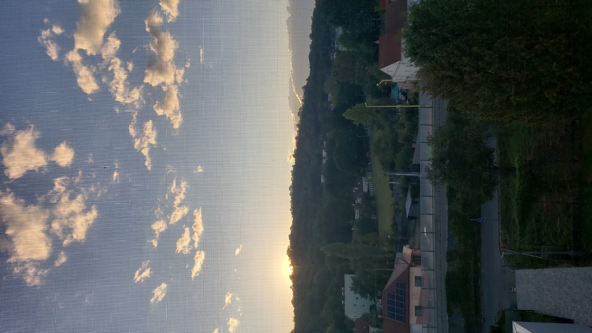 This is a vertical, medium-shot, outdoor photograph taken at dusk in Prešov, Slovakia. The image is captured through a window screen, which creates a grid pattern across the scene. The sky dominates the upper portion of the frame, filled with scattered, illuminated clouds against a pale blue backdrop.
In the middle ground, a tree-covered hill forms a dark, undulating line against the horizon. The setting sun is visible as a bright, hazy orb peeking from behind the crest of the hill on the left, casting a warm glow. Below the hill, a suburban landscape unfolds, featuring several buildings with red and white roofs, some of which appear to be houses. Solar panels are visible on the roof of a building in the lower left quadrant. A road or street runs horizontally across the middle of the image, bordered by greenery and a fence.
The foreground consists of a dark, shadowed area with some paving stones visible in the lower left, suggesting a balcony or walkway. The overall impression is a peaceful, residential view at the end of the day.