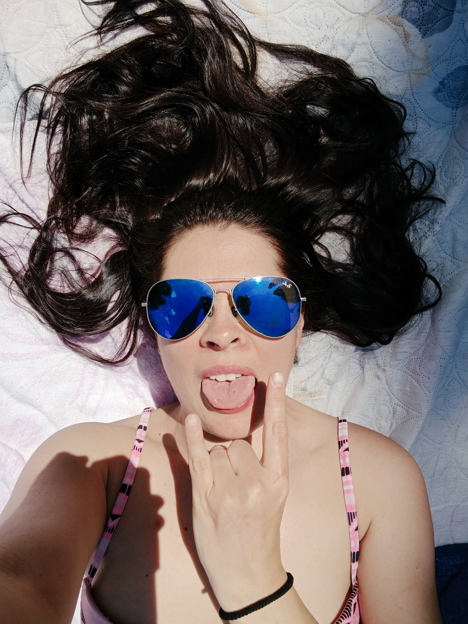 A person lies on their back on a white blanket, with their head to the right. The person has long, dark, wavy hair spread out around their head and shoulders. They are wearing aviator sunglasses with bright blue mirrored lenses, which reflect the sky and a hand holding a phone. The person's mouth is open, with their tongue sticking out, and their right hand is raised, making a rock-and-roll sign with their index and pinky fingers extended. The person is also wearing a pink and white patterned bikini top. The lighting suggests they are outdoors, possibly in a sunny location.