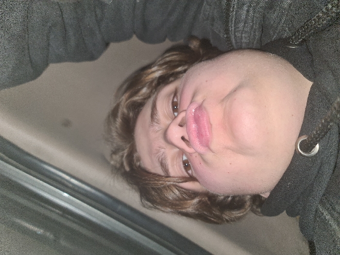 A close-up selfie features a person with light skin, brown eyes, and light brown curly hair. They are wearing a dark gray hooded sweatshirt with the drawstring visible. The camera angle is tilted, causing a distorted perspective, particularly noticeable in the stretched facial features and pursed lips. A gray surface is partially visible on the left side of the frame. The overall lighting is dim.