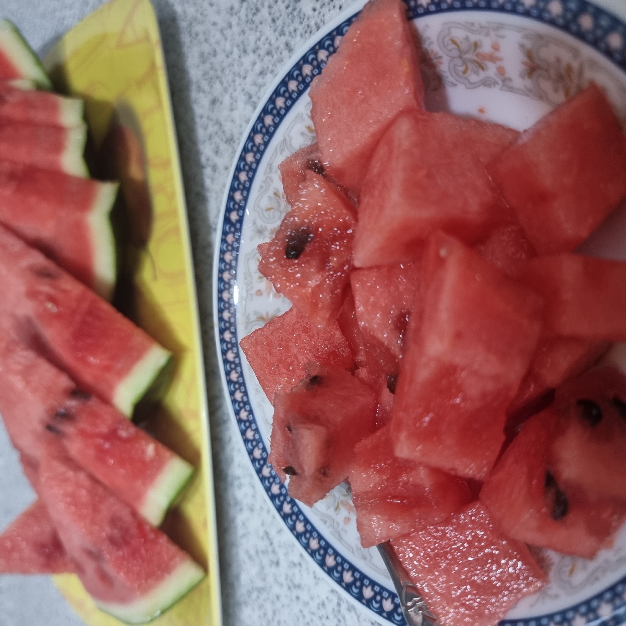 This media file shows a close-up, overhead view of a plate of sliced watermelon and a yellow tray also filled with watermelon slices. The watermelon is cut into bite-sized cubes and irregular triangular pieces. Some pieces still have black seeds visible. The watermelon is a vibrant red color with a thin white rind and a green outer peel on some slices.
The watermelon slices are arranged on two surfaces. The left side of the image features a yellow tray with several triangular slices of watermelon. The right side of the image displays a white plate with a decorative blue and white patterned rim, filled with cubes and larger chunks of watermelon. A silver fork is partially visible on the plate, suggesting the watermelon is ready to be eaten.
The background appears to be a speckled countertop, possibly in a kitchen or dining area. The lighting suggests it is daytime, likely indoors. There is no visible text in the image. The scene depicts a simple, refreshing snack, likely prepared for enjoyment. The context of "Dunnae-myeon, South Korea" suggests a domestic setting in this region.