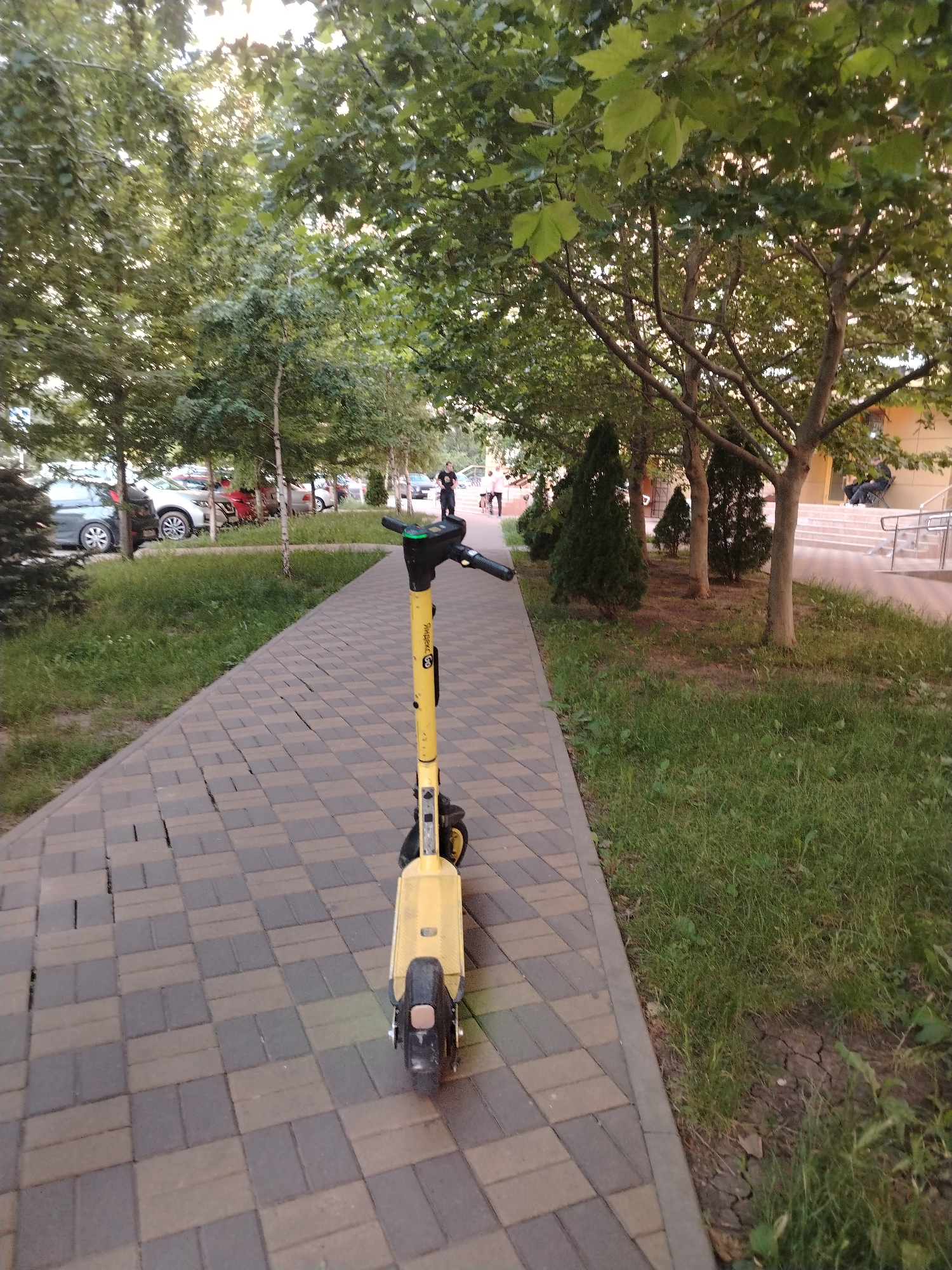 A yellow electric scooter is parked on a paved walkway in a residential area of Krasnodar, Russia. The walkway is made of interlocking rectangular stones in shades of brown and gray. To the left of the walkway is a strip of grass, and beyond that are parked cars. To the right of the walkway is a grassy area with trees and a building in the background. A person wearing a dark shirt and light-colored shorts is walking away from the camera on the walkway. The sky is visible through the leaves of the trees.