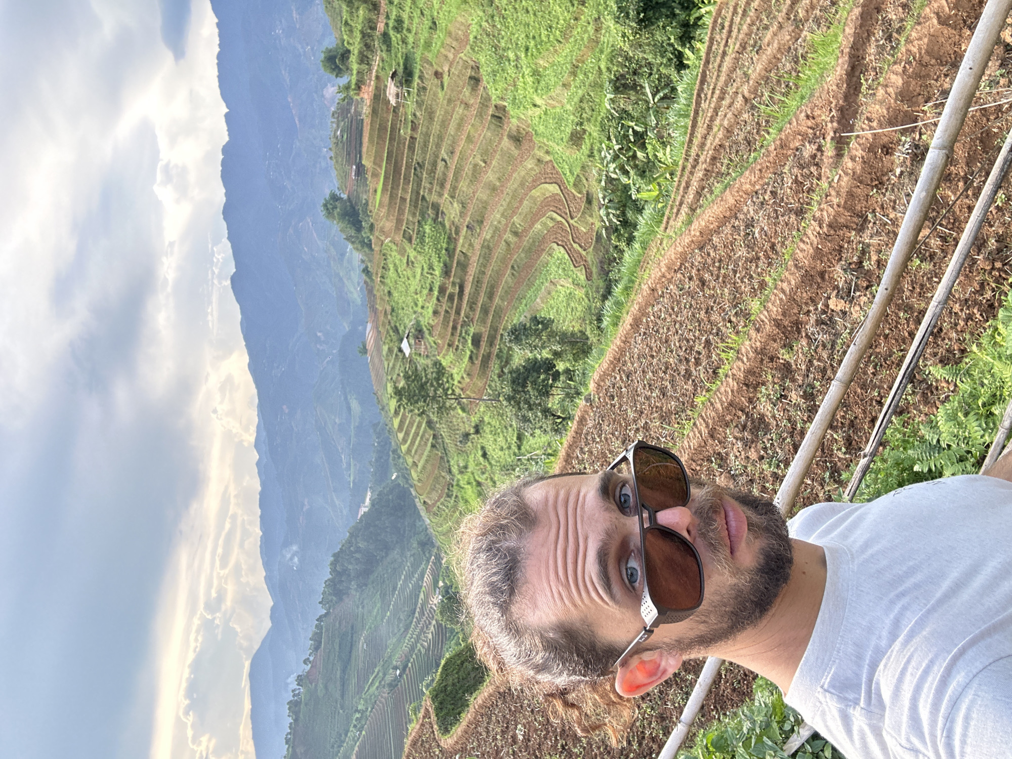 A horizontal outdoor shot captures a man posing in front of a vast mountainous landscape characterized by agricultural terraced fields, likely in Lao Chai, Vietnam.
In the foreground, a man with light brown hair, a beard, and blue eyes looks directly at the camera. He is wearing sunglasses with dark brown lenses and a white t-shirt. His brow is slightly furrowed. A part of a railing or fence made of light-colored poles is visible to his right, suggesting he is standing on a viewpoint or platform.
The background showcases impressive terraced fields that cover the hillsides. Some terraces are lush green with vegetation, while others appear as exposed brown earth, indicating freshly tilled soil or different stages of cultivation. Small, simple structures, possibly huts or shelters, are dotted amongst the terraces. Beyond these cultivated slopes, majestic mountains rise, appearing a muted blue under a largely cloudy sky. Patches of brighter, whiter clouds are interspersed with darker grey ones, suggesting a partly cloudy or overcast day. The light is bright and even, indicative of daytime.
The image presents a blend of human presence within a grand natural and cultivated environment. No visible text is present in the image.