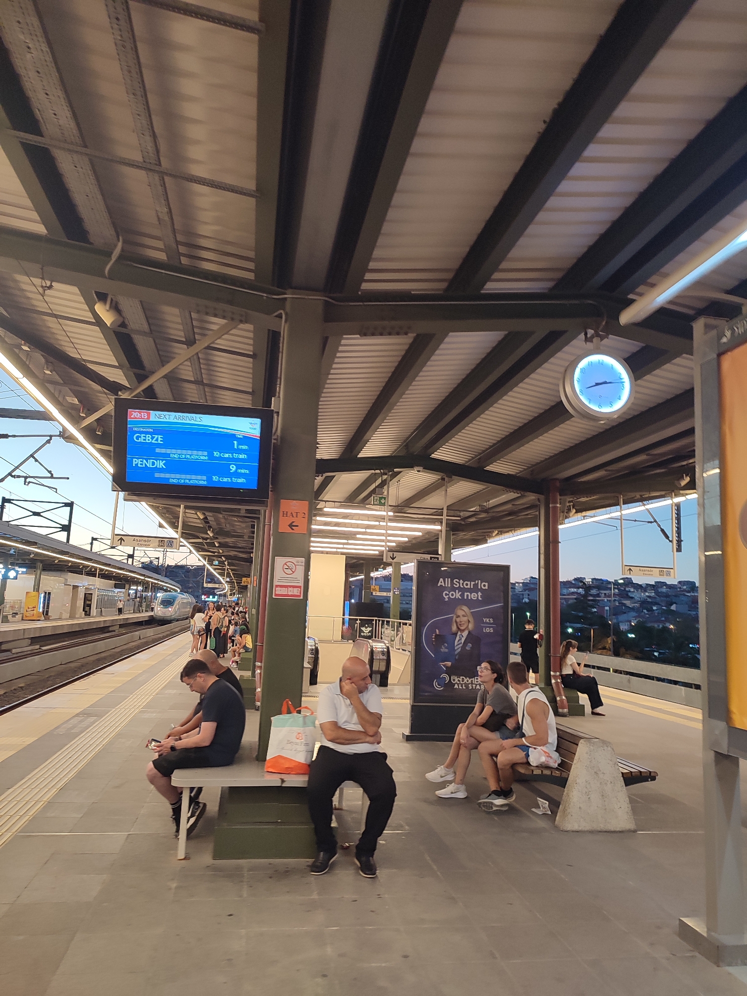 The image depicts a train station platform in Kadıköy, Turkey. In the foreground, several people are seated on benches. On the left, a man in a black t-shirt and shorts is looking at his phone. Next to him, a man in a white t-shirt and black pants is resting his head on his hand. Further down the platform, a couple is seated closely together on a bench; the woman is wearing a grey t-shirt and shorts, and the man is in a white tank top and blue shorts. Behind them, a woman in a white top and black pants is sitting on another bench.
Above the platform, a large digital display shows train arrival information, with "GEBZE" listed as a destination with an arrival in 1 minute and "PENDIK" listed with an arrival in 9 minutes. A clock is visible on the ceiling structure.
In the background, a modern train is stopped at the platform. Other passengers are visible walking and waiting. The station features a covered roof supported by metal pillars. The cityscape is visible in the distance, with buildings nestled on a hillside under a twilight sky. Advertisements are displayed on posters, including one for "All Star'la çok net". Signs indicate "HAT 2" for Platform 2. The overall environment suggests a functional and somewhat crowded public transportation hub during the evening.
