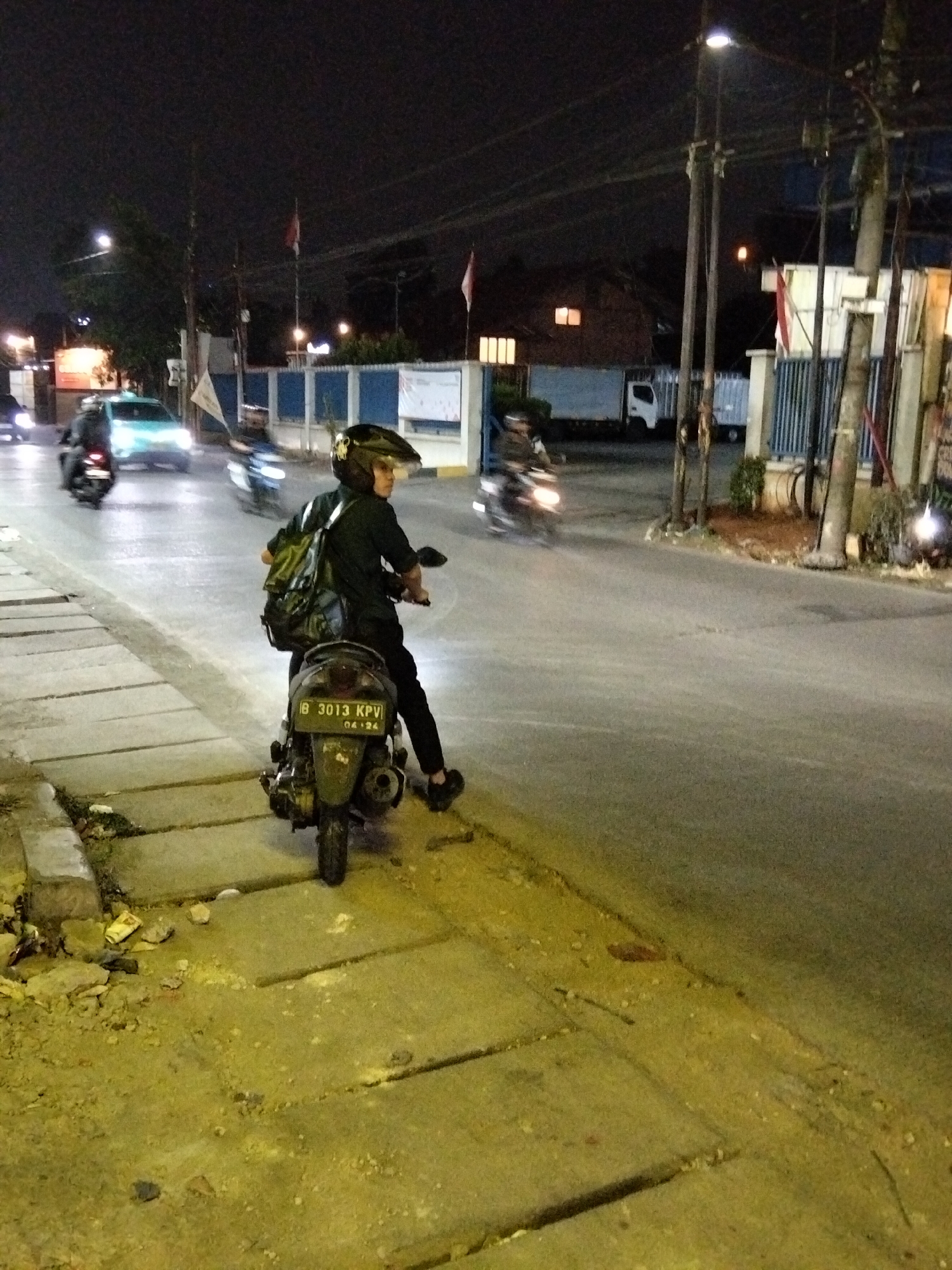 A person wearing a black helmet, black shirt, and black pants rides a dark colored scooter at night on a paved road in South Tangerang, Indonesia. The scooter has a license plate that reads "B 3013 KPV 04-24". The person is carrying a green backpack. The scooter is parked on the side of the road, with its rear facing the camera. The road is lit by streetlights. In the background, there are buildings, trees, and other vehicles. The sidewalk on the left side of the image is made of concrete slabs and is covered with debris. The overall scene depicts an urban environment at night.