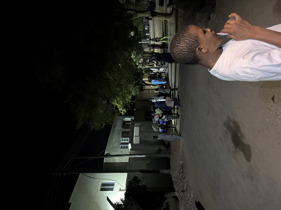 The scene depicts a nighttime street in Gandu Albasa, Nigeria. In the immediate foreground, to the right, a young male with short, dark hair, wearing a white t-shirt, is visible from the chest up, facing left. He is holding a white object, consistent with a toothbrush, to his mouth, suggesting oral hygiene. The ground directly in front of him is an unpaved dirt surface with a dark, irregular liquid stain.
In the midground, an unpaved road extends into the distance, illuminated by artificial light sources. On the left side of the road, a pile of dark debris or rubble is present. Beyond this, a two-story building with a light-colored, possibly white or cream, facade and a visible window is illuminated. Adjacent to it, a utility pole with overhead wires stands. To the right of the pole, another building with a grey facade is visible, featuring a horizontal-slatted metal gate and two windows with white frames on its upper story.
A group of approximately 15-20 individuals, predominantly adult males, are gathered on the road between the buildings and trees. Many are dressed in traditional robes of various colors, including blue, grey, and white. A blue vehicle is partially obscured by the group. Most individuals are standing, appearing to engage in casual conversation or observation. One person on the far left of the group wears a light grey robe. Further down the street, to the right, a person is seen with a bicycle.
Trees with visible foliage line the right side of the street and extend into the background, with some leaves illuminated by ambient light. The sky is dark, indicating night. Faint lights from distant buildings or streetlights are visible further down the road, contributing to the overall illumination of the scene.