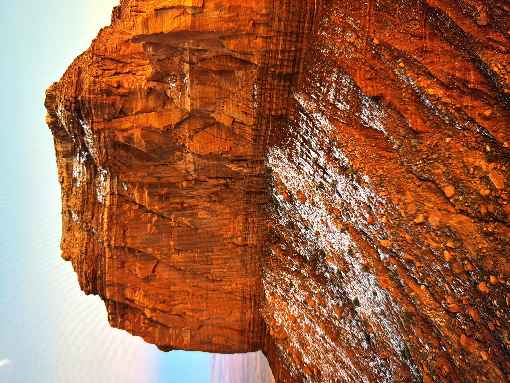 This is an image of a large, stratified rock formation with a reddish-orange hue, possibly sandstone. The rock face is rugged and shows distinct horizontal layers. The lower part of the formation is covered in a sparse layer of white snow and scattered rocks. The sky above is a pale blue, transitioning to a lighter hue towards the horizon. The overall scene depicts a natural landscape, likely a desert or canyon environment. This image is not located in the city of Kyiv, Ukraine.