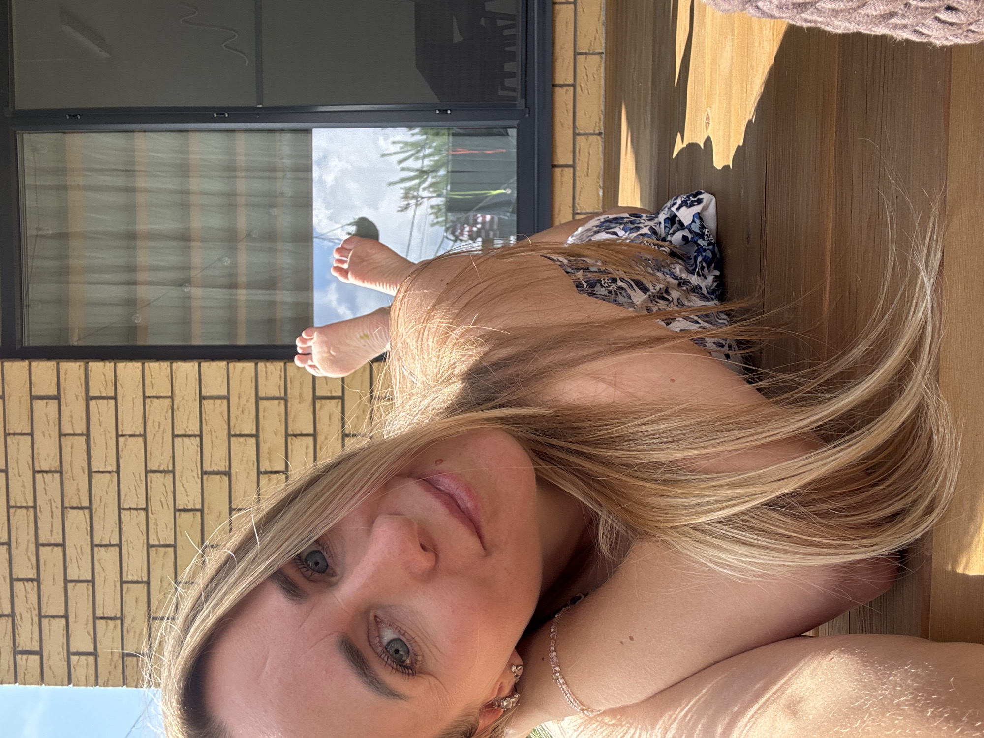 A woman with blonde hair is lying on a wooden deck, resting her head on her arms. She is wearing a patterned swimsuit. Her feet are visible in the background, propped up. The scene is outdoors, with a brick wall and a window behind her. The location is not specified in the image and cannot be determined to be Chaikovsky, Russia.