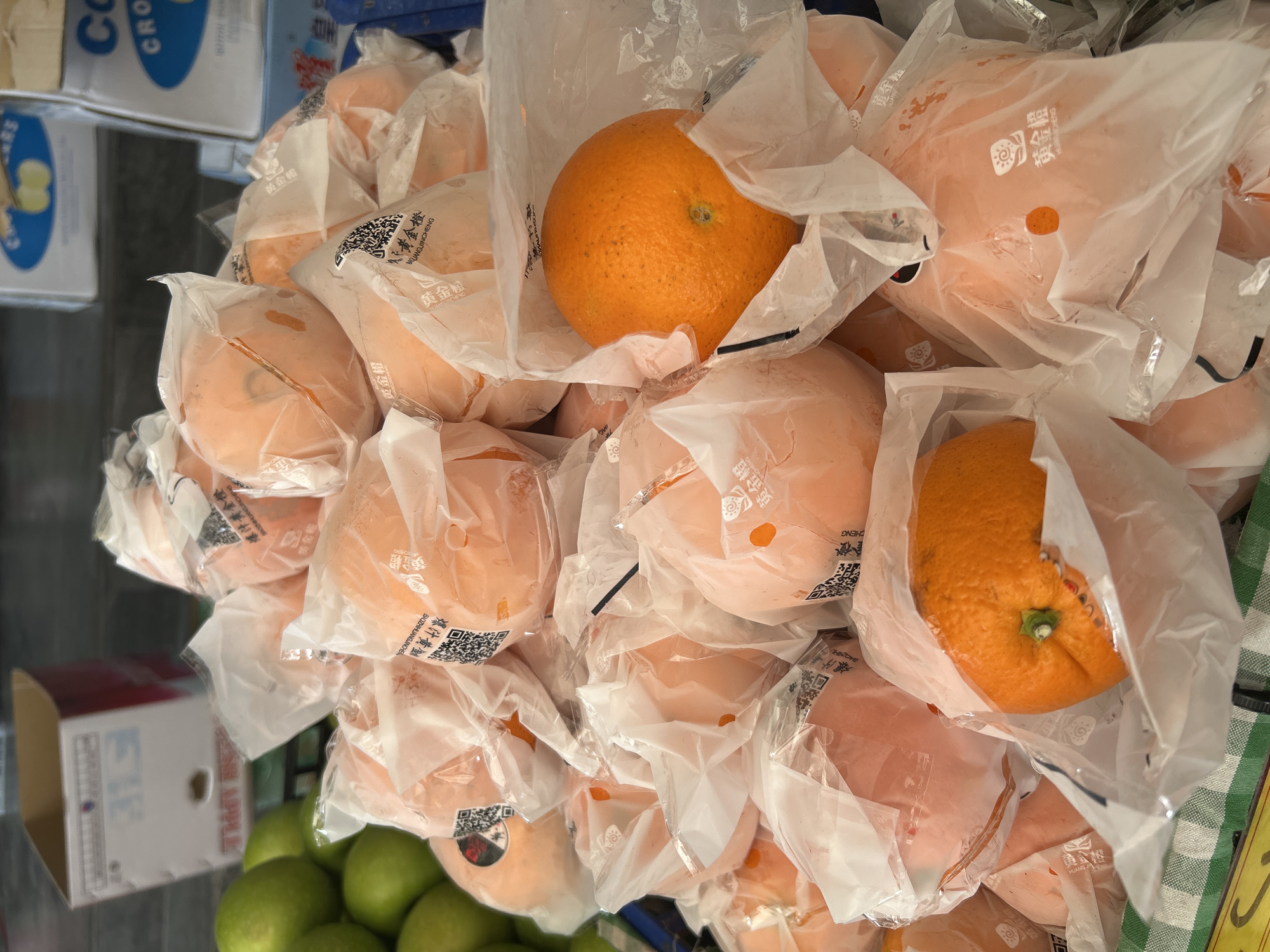 The image shows a close-up, top-down view of a large pile of oranges, many of which are individually wrapped in clear plastic bags. Some of the oranges are whole and have visible texture, while others appear softer and possibly juiced or pureed. The oranges are predominantly orange in color, with a few lighter, peachy-toned ones also present.
The background reveals glimpses of a market stall setting. To the left, there are boxes with text in what appears to be Chinese characters, some of which are partially obscured by the fruit. The text "COS" and "CRO" are visible on one of the boxes, along with branding that suggests apples ("FRESH APPLE"). Below the oranges, a cluster of green apples is visible. The overall lighting suggests an indoor or shaded environment.
Several of the plastic bags containing the oranges have text printed on them, including Chinese characters and what looks like branding. The phrase "黄金橙" (Huángjīn chéng), meaning "golden orange," appears repeatedly. Other text includes "爆汁" (bào zhī), meaning "bursting with juice," and partial English words like "JUICE" and "APPLE."
The image is an object-focused shot of produce for sale, likely in a market in Sukawati, Indonesia. There are no people visible, and the focus is entirely on the arrangement and packaging of the fruit. The visual suggests a commercial display of fresh produce.