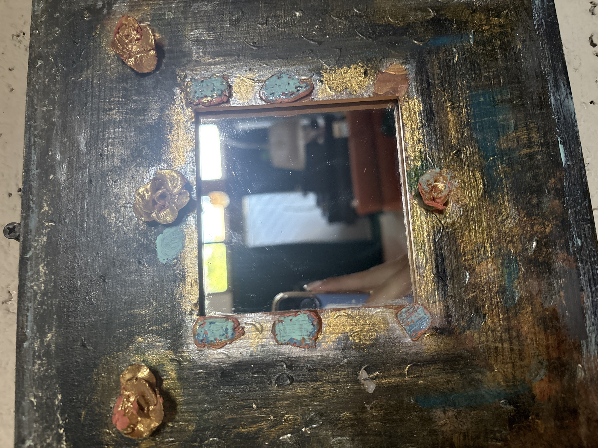 This is a close-up, eye-level, indoor shot of a square, ornate mirror. The mirror is framed by a dark, textured material with gold and teal paint accents. Small, three-dimensional embellishments, appearing to be made of textured material with gold and pink coloring, are attached around the frame. Some of these embellishments resemble roses. Small, chipped teal-colored accents with a red undertone are also present on the frame, surrounding the mirror. The mirror itself reflects a room with bright lighting, furniture, and a hand holding a phone. The background of the overall image is a light-colored, textured wall with a screw visible in the upper right corner. The scene is consistent with an indoor decorative object in a residential or studio setting.