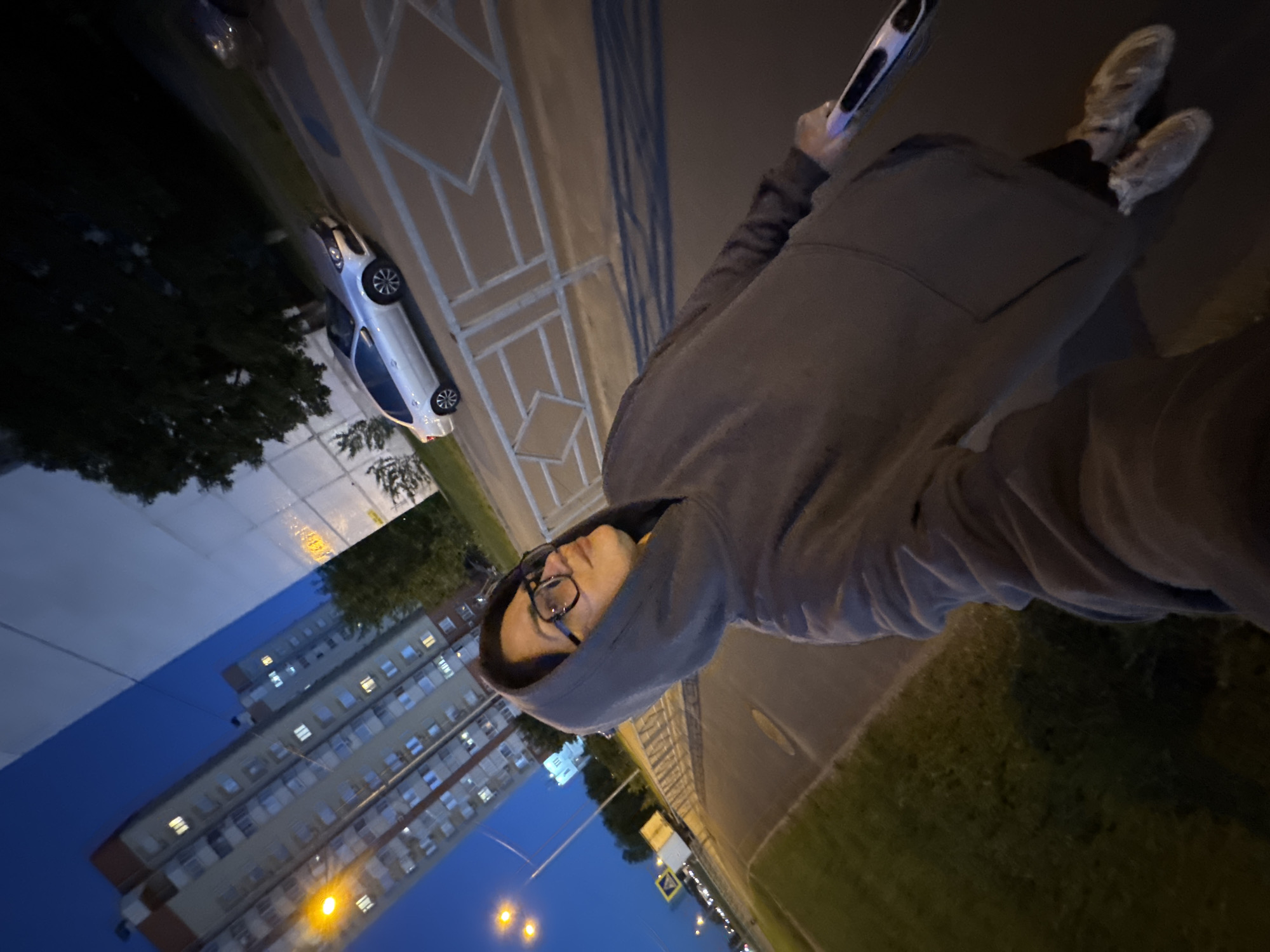 A person wearing glasses and a gray hooded sweatshirt holds a black-and-white device in their hand, standing on an urban sidewalk. The street features a white car parked along the curb, and buildings with illuminated windows are visible in the background under a blue evening sky. Streetlights and road signs dot the landscape. Adjacent to the sidewalk is a green, grassy area.